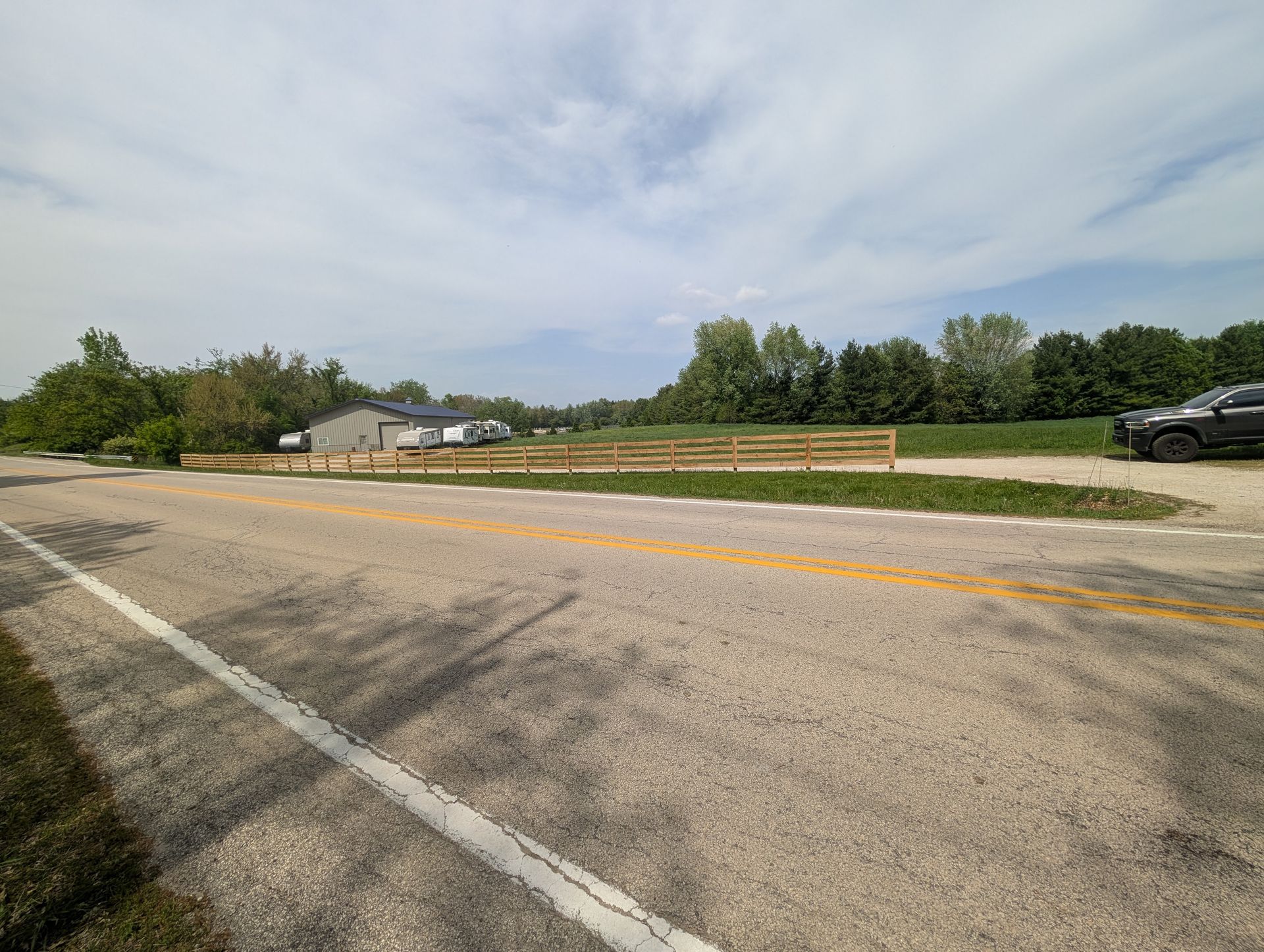 A rural road stretches into the distance, with a wooden fence bordering a field, a house, and trees under a blue sky.