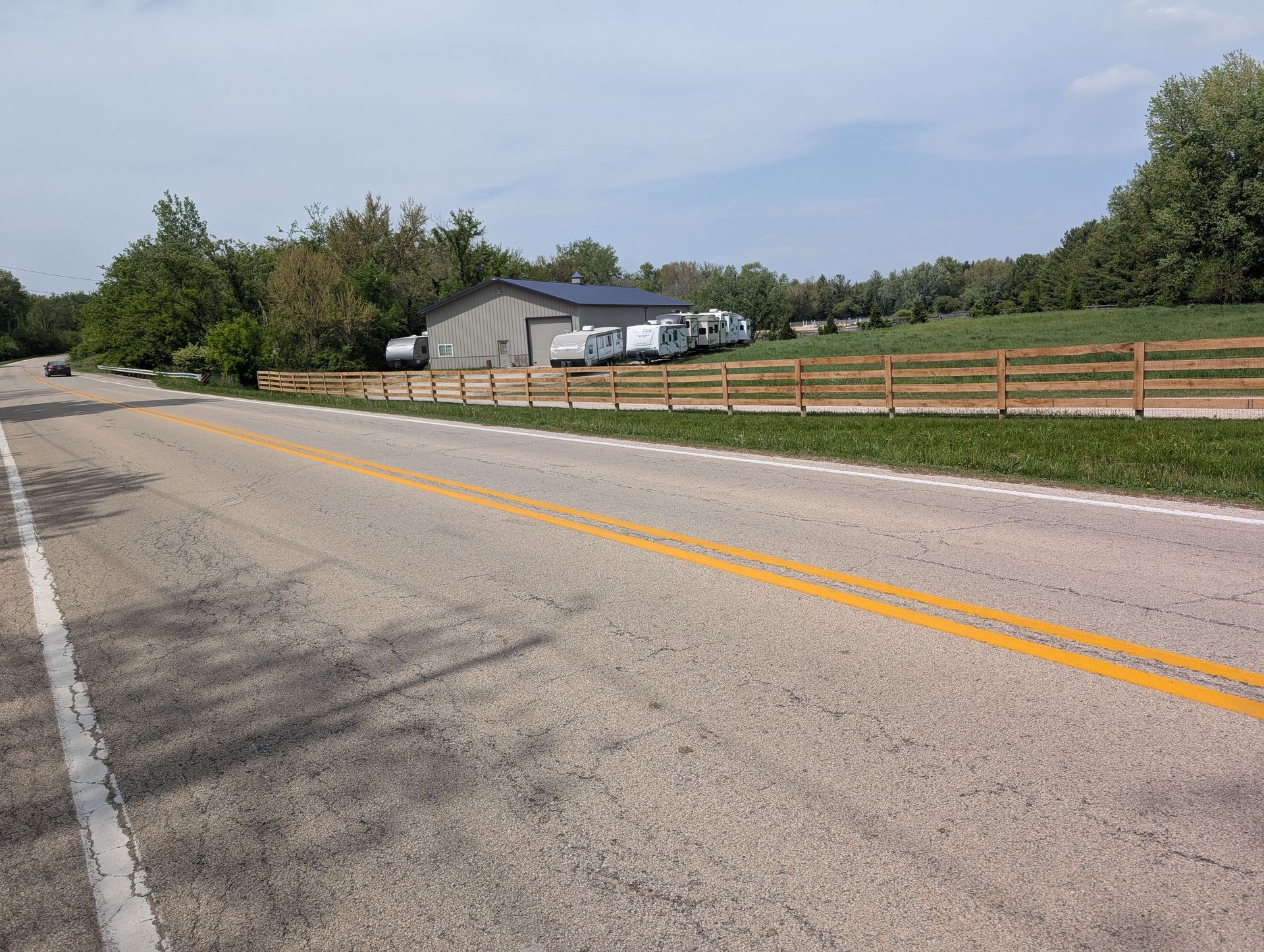A rural road stretches past a wooden fence and a metal warehouse where several white travel trailers are parked.