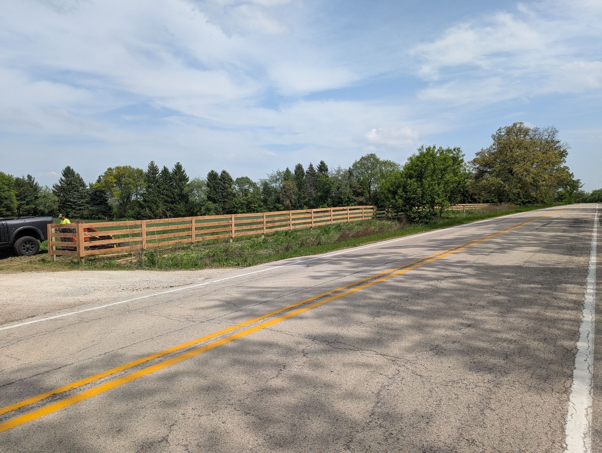 A wooden post-and-rail fence lines a gravel road beside a dense green forest under a blue sky with light clouds.