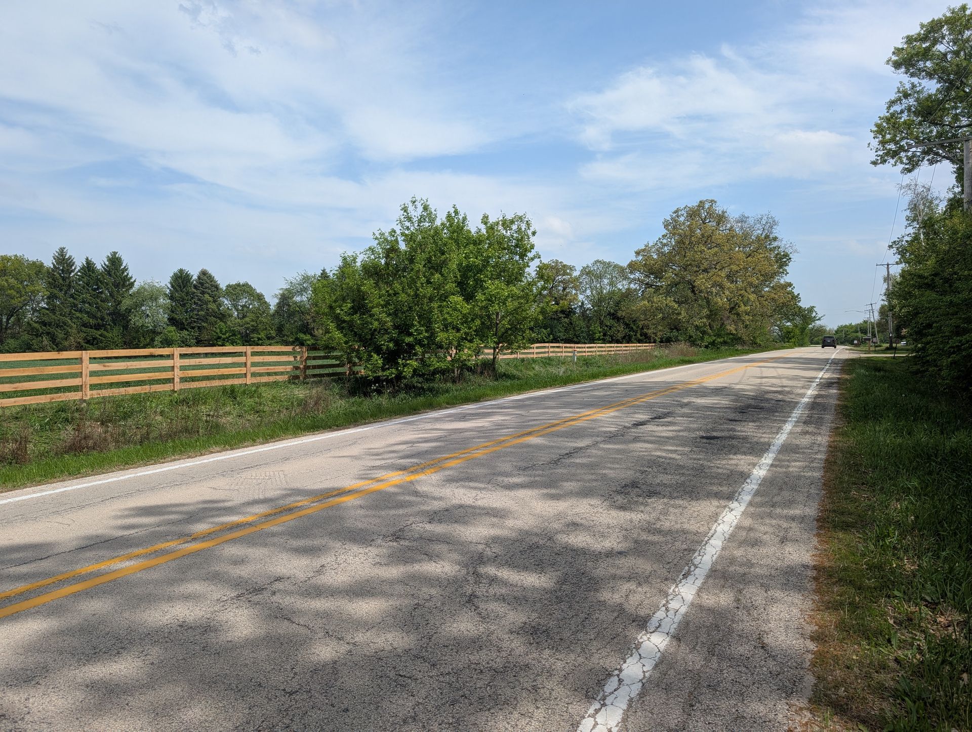 A rural asphalt road stretches into the distance under a blue sky, bordered by a wooden fence and lush green trees.