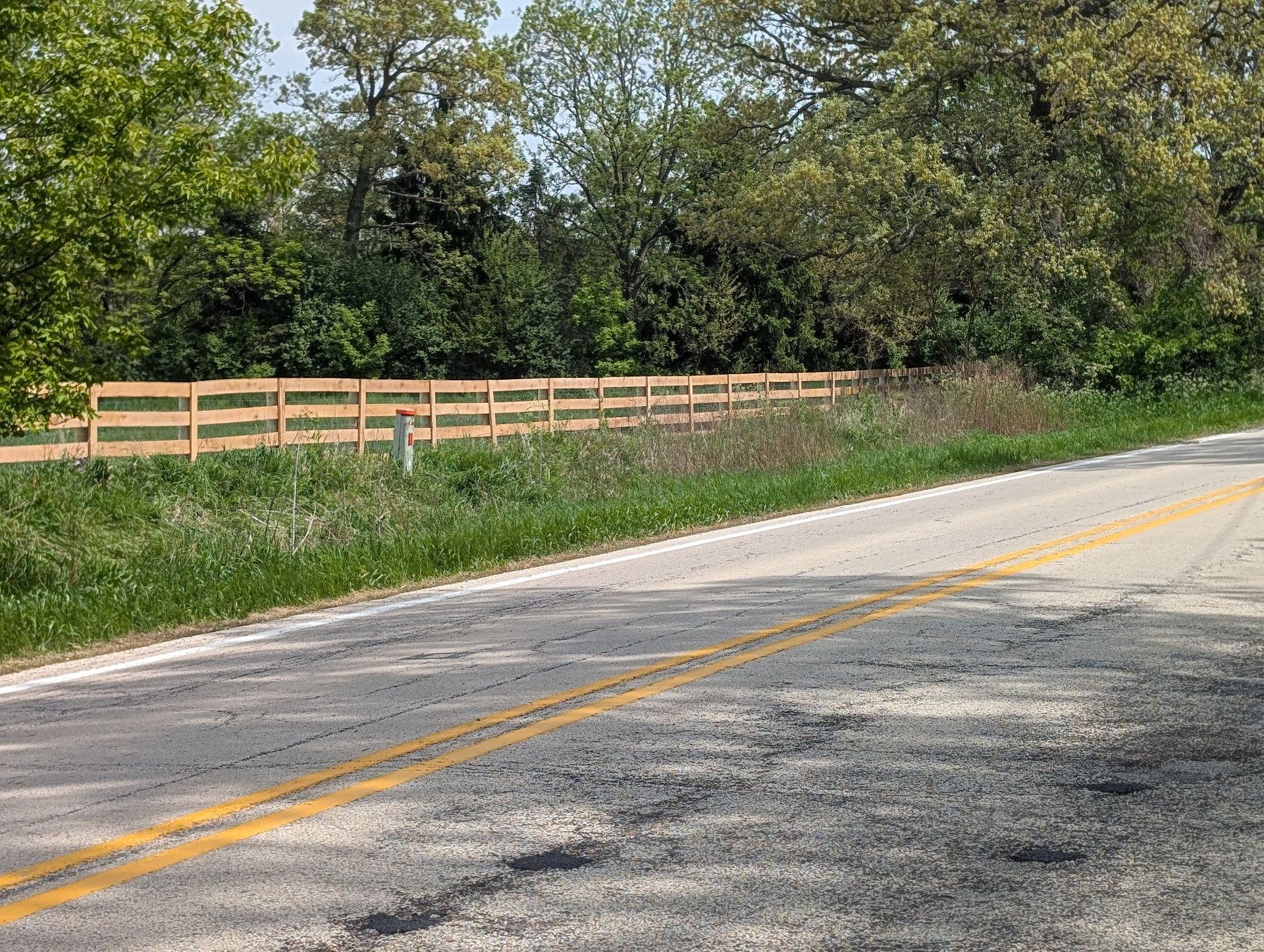 A paved country road with a double yellow line running beside a grassy verge and a light-colored wooden rail fence.