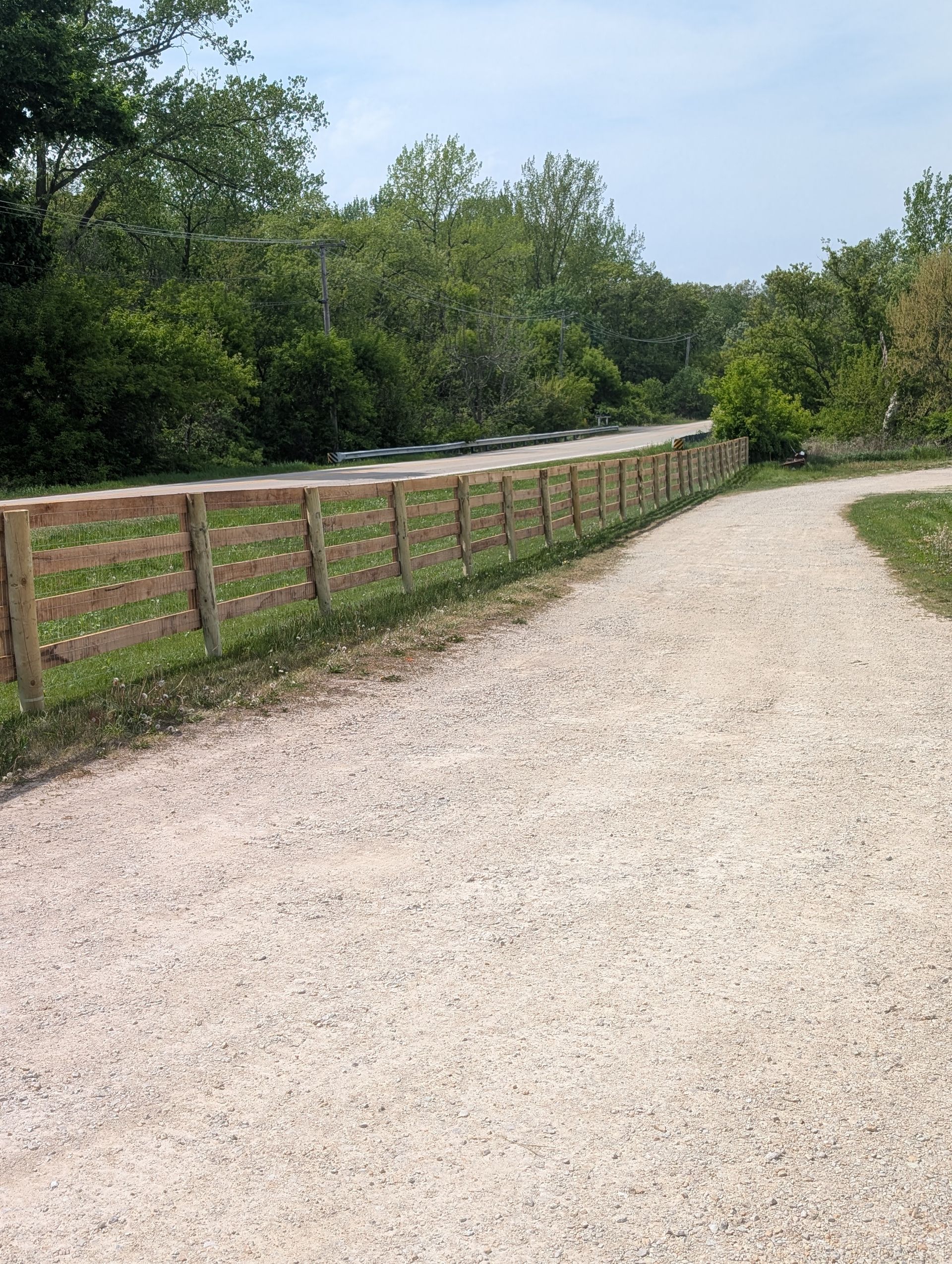 A gravel path leads toward a wooden split-rail fence alongside a paved road, bordered by dense green trees under a blue sky.