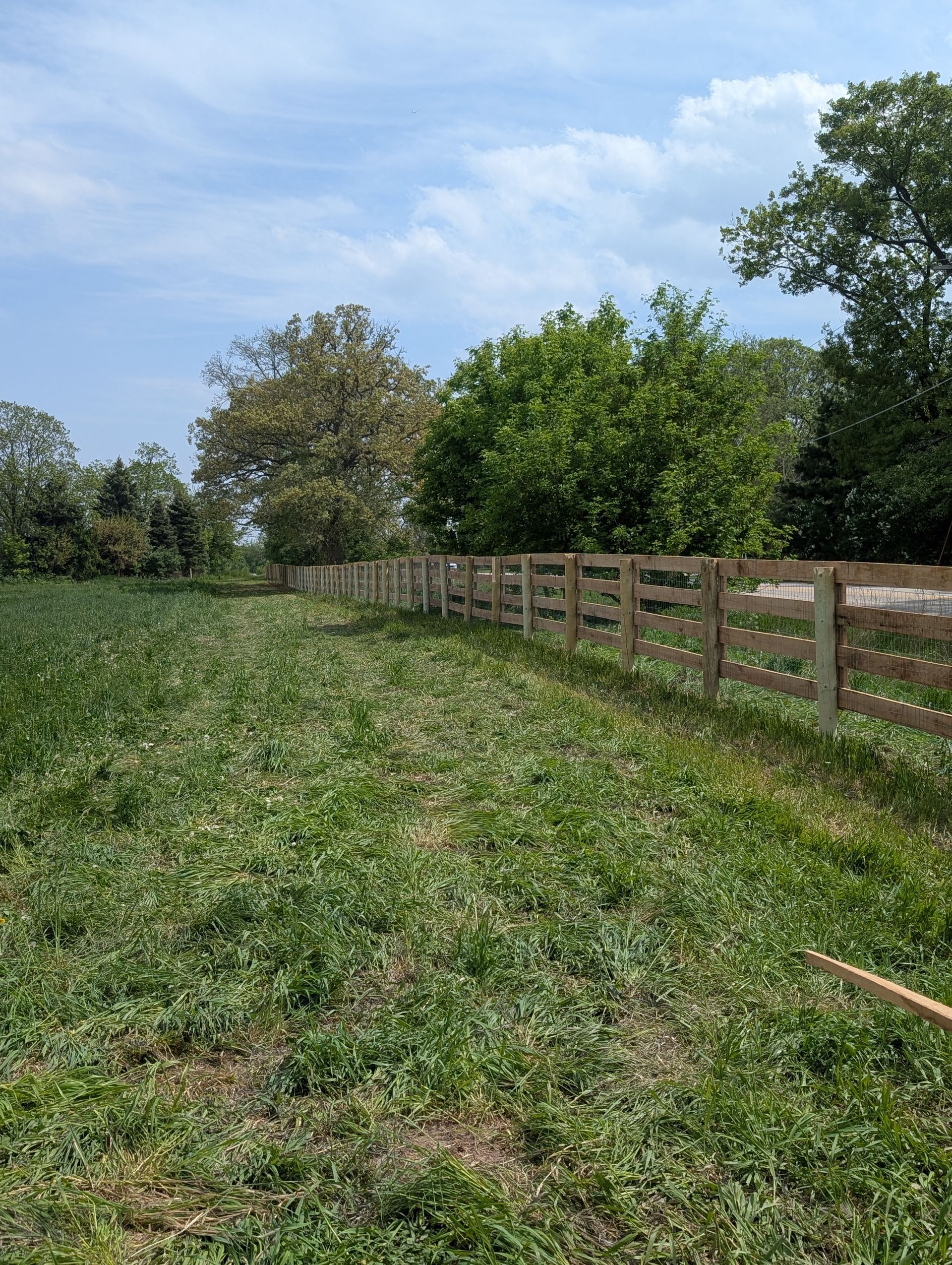 A wooden post-and-rail fence runs alongside a green grassy field under a bright, partly cloudy blue sky.