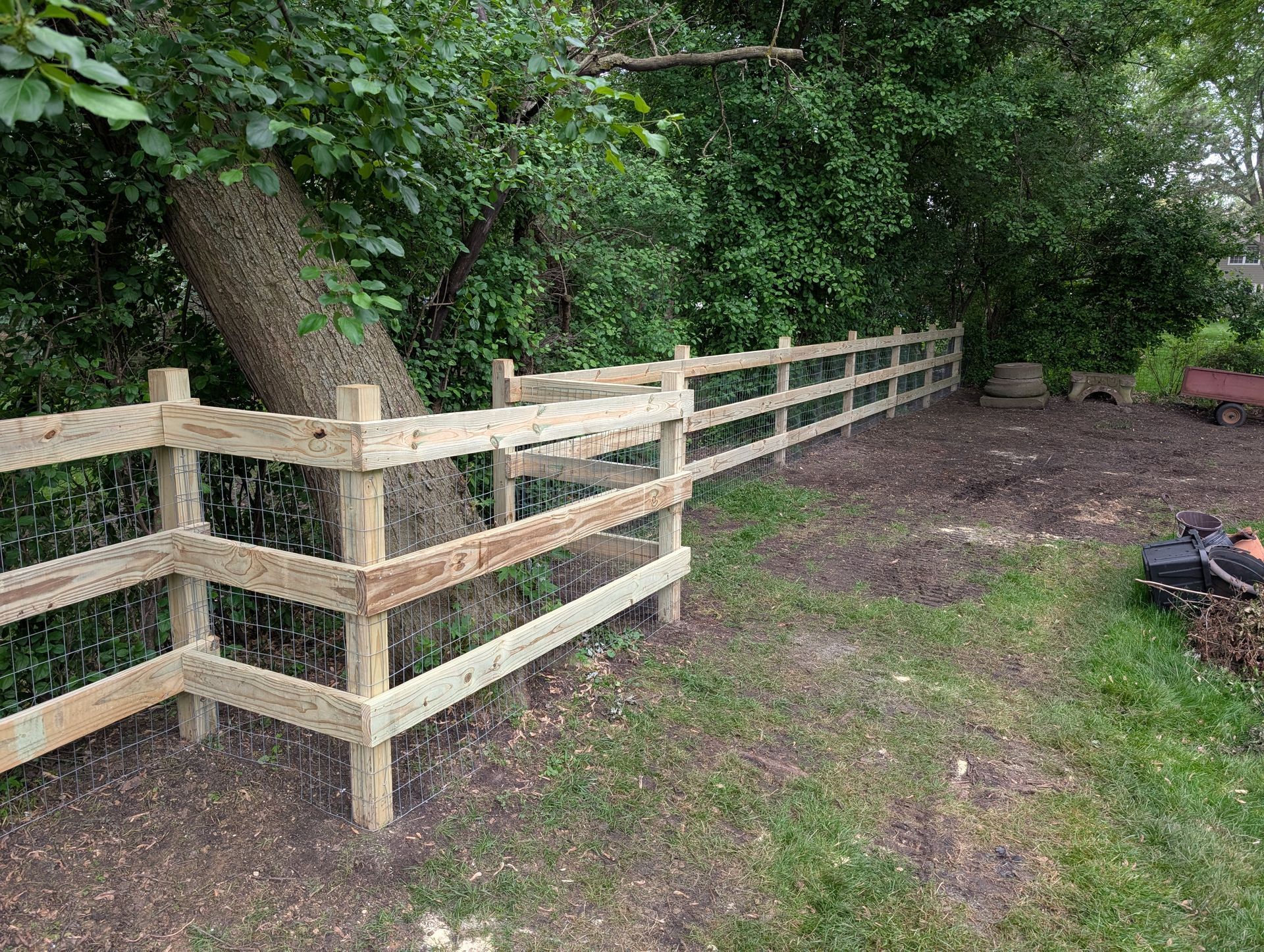 A new three-rail wooden fence spans a grassy yard, partially wrapping around a large tree near a wooded area.