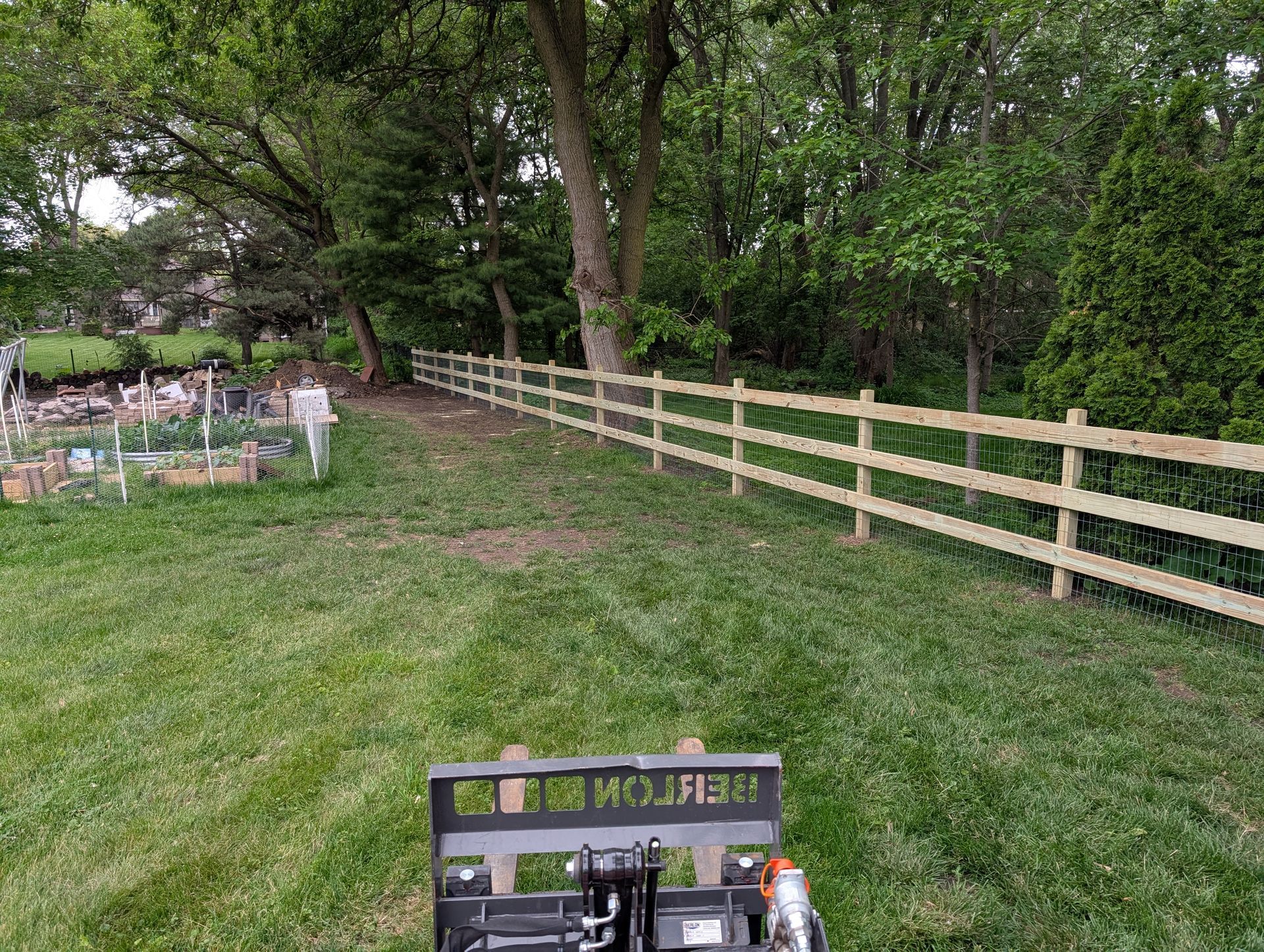A three-rail wooden fence borders a green lawn and wooded area, viewed from behind a piece of machinery.