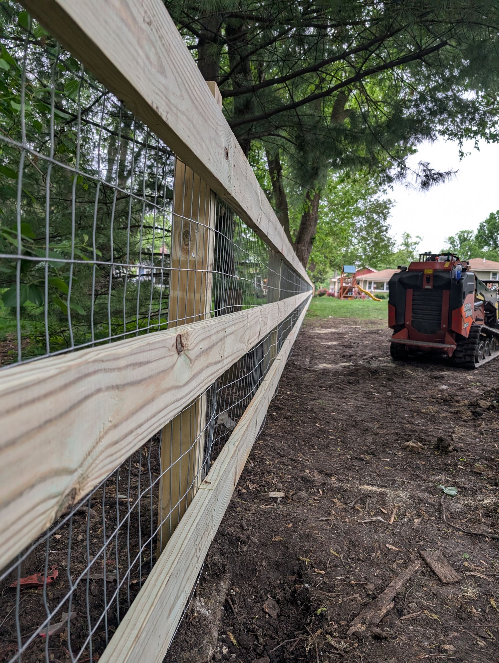 A low-angle view of a newly installed wooden fence with wire mesh, featuring a construction vehicle parked in the distance.