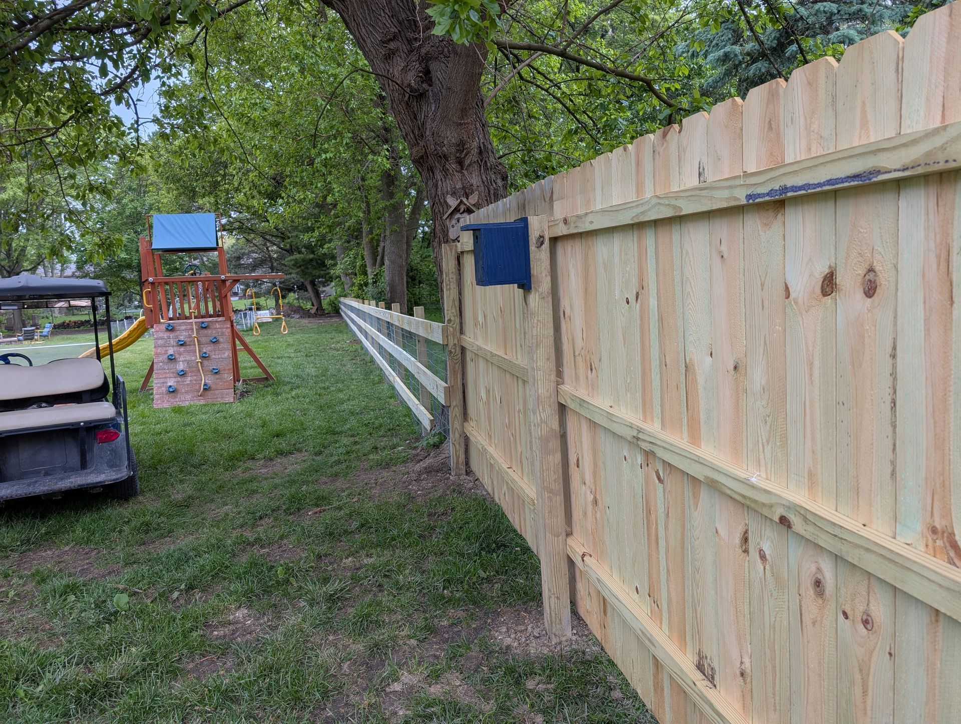 A wooden fence in a grassy yard near a small children's playground set and a golf cart.