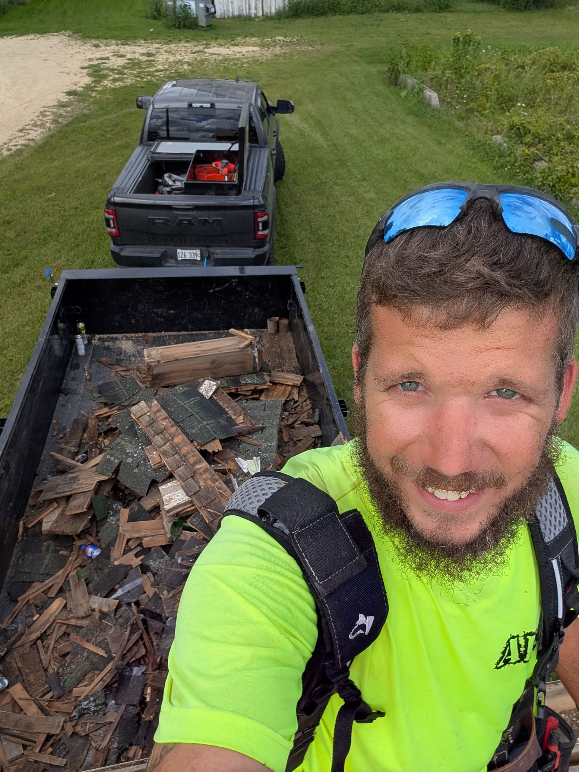 A worker in a bright yellow shirt smiles for a selfie in front of a pickup truck pulling a trailer full of roof shingles.
