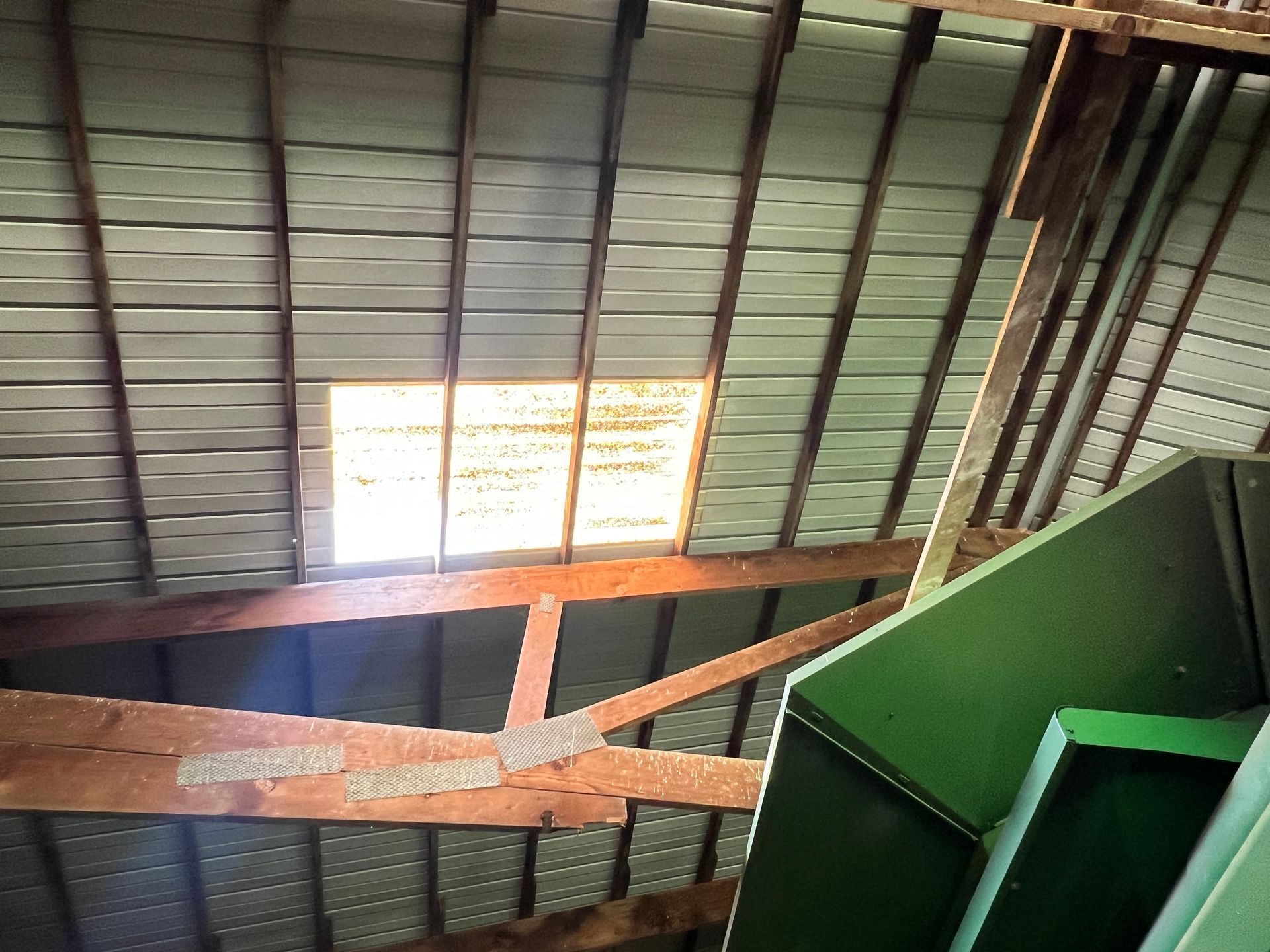 An interior view of a barn roof featuring dark wooden beams, metal roofing panels, and a large opening letting in light.