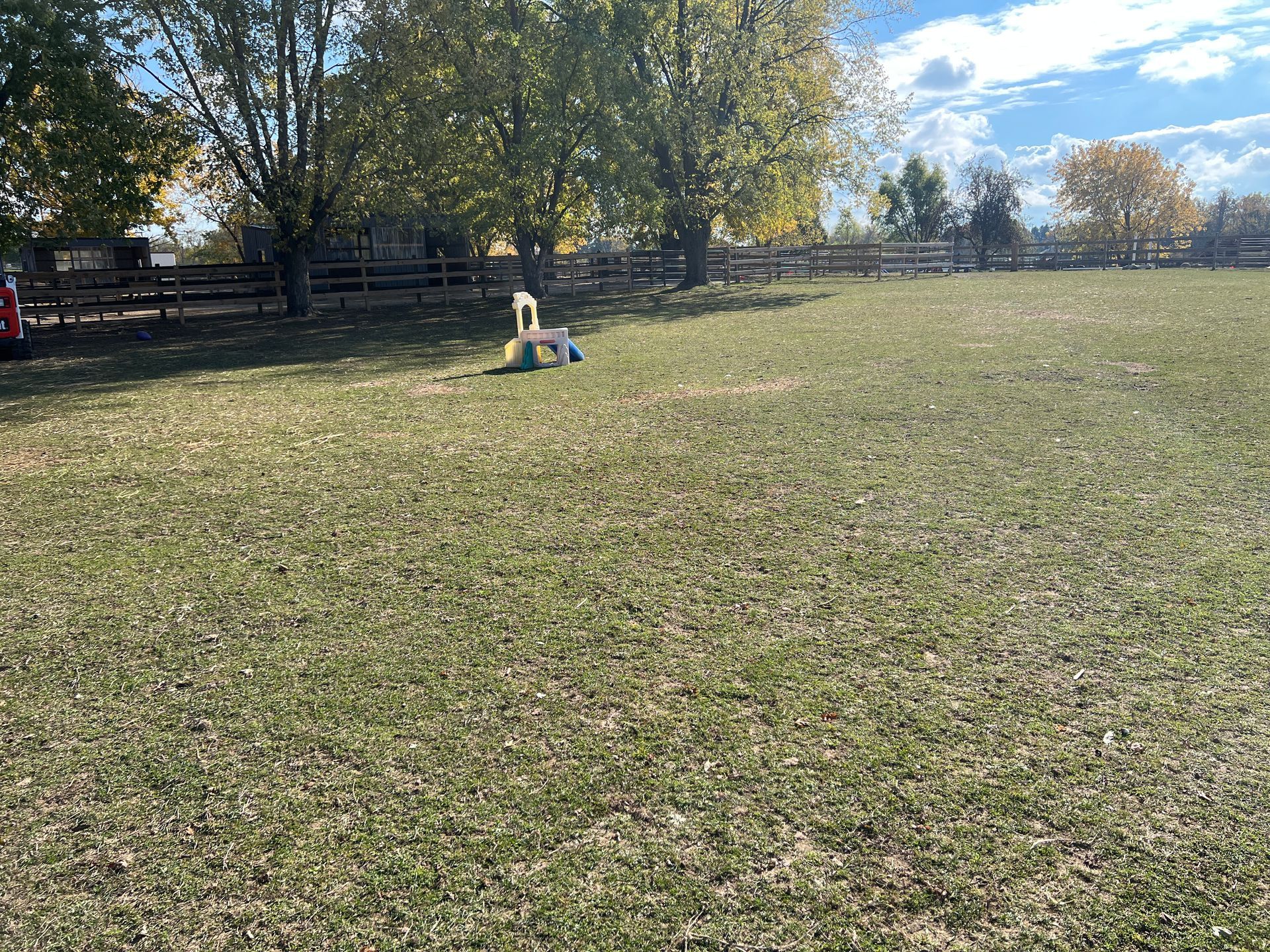 A lone white plastic chair stands in the middle of a large, grassy field near a wooden fence and trees under a sunny sky.
