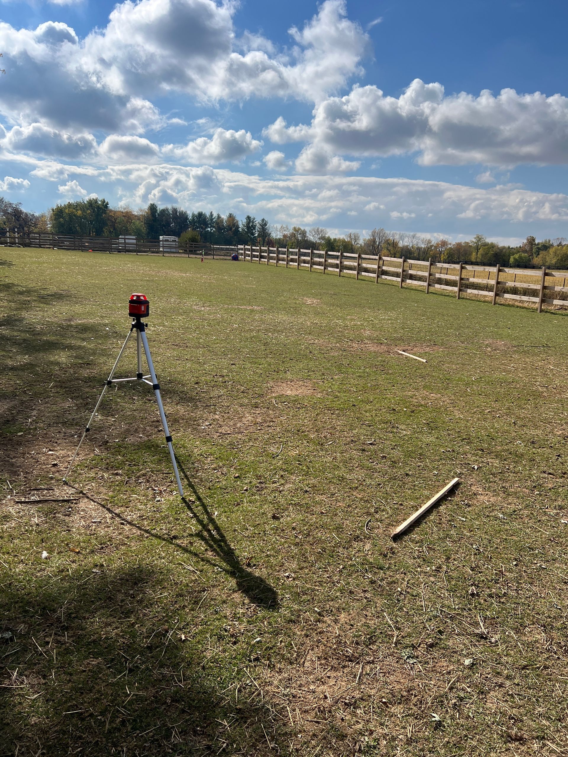 A red rotating laser level on a tripod stands in a grassy field under a bright, partly cloudy blue sky.