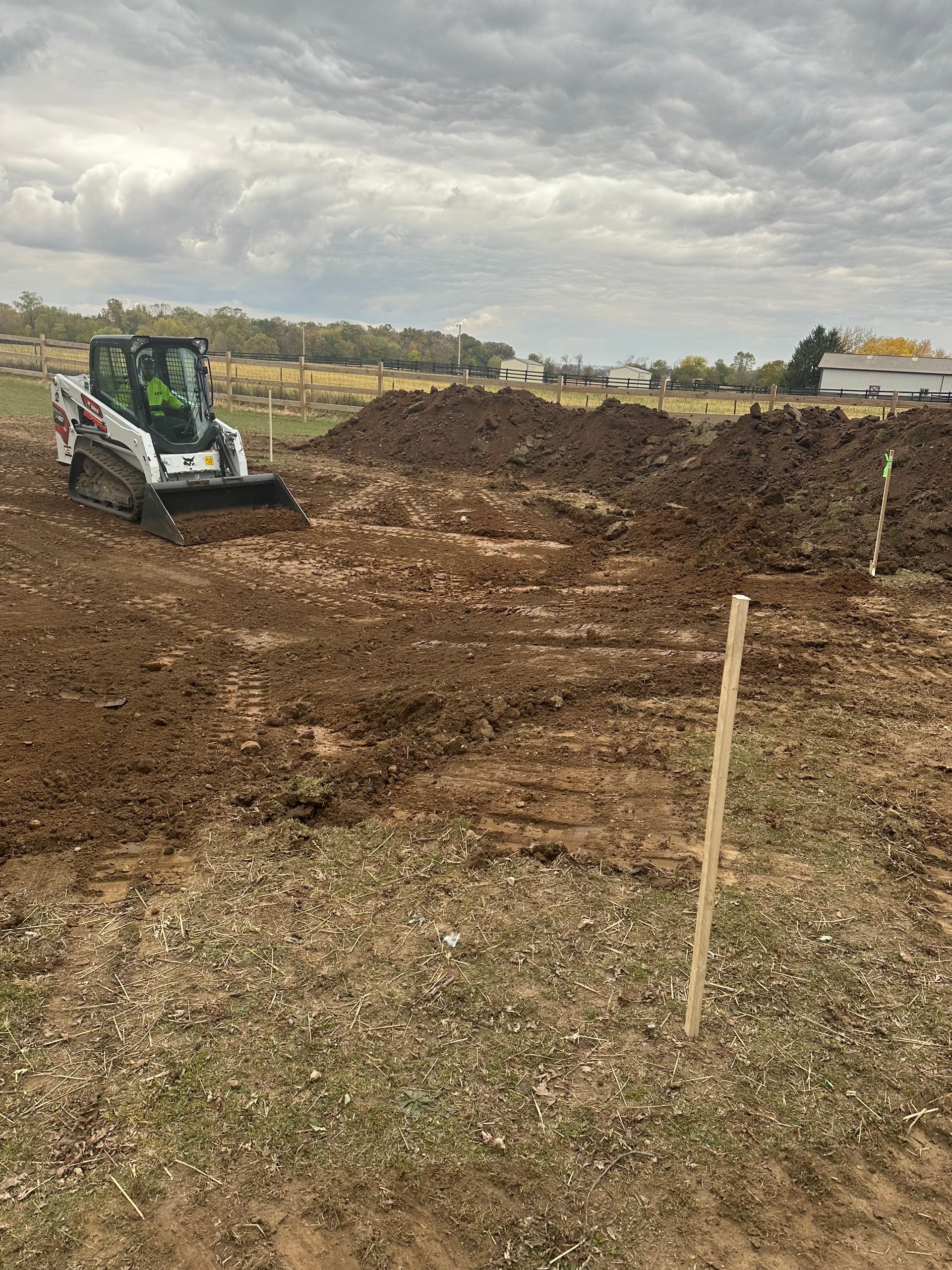 A white skid-steer loader works in a dirt field under a cloudy sky, near large mounds of excavated soil and a survey stake.
