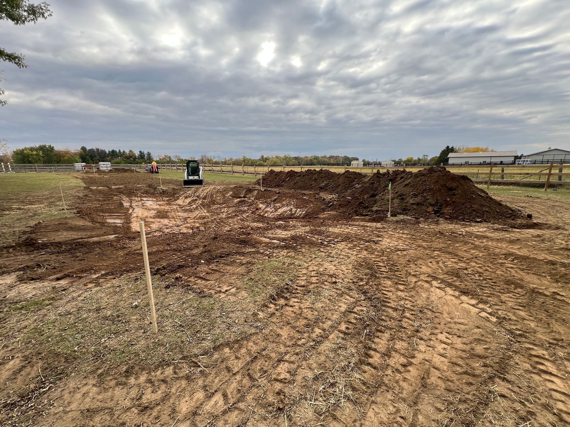 A construction site with a small excavator parked near a large pile of excavated dirt under a cloudy sky.