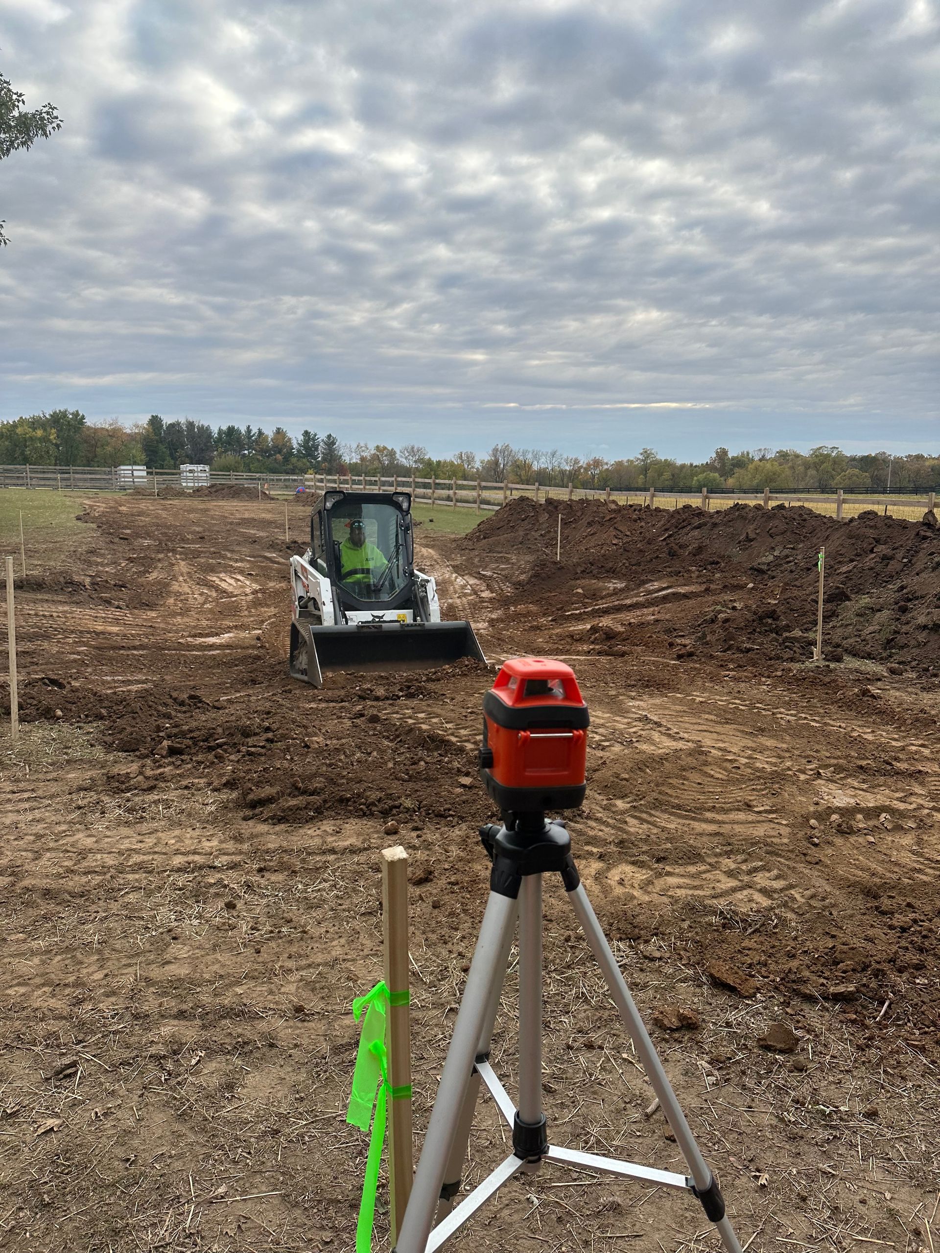A red laser level on a tripod faces a white skid steer working on a dirt construction site under a cloudy sky.
