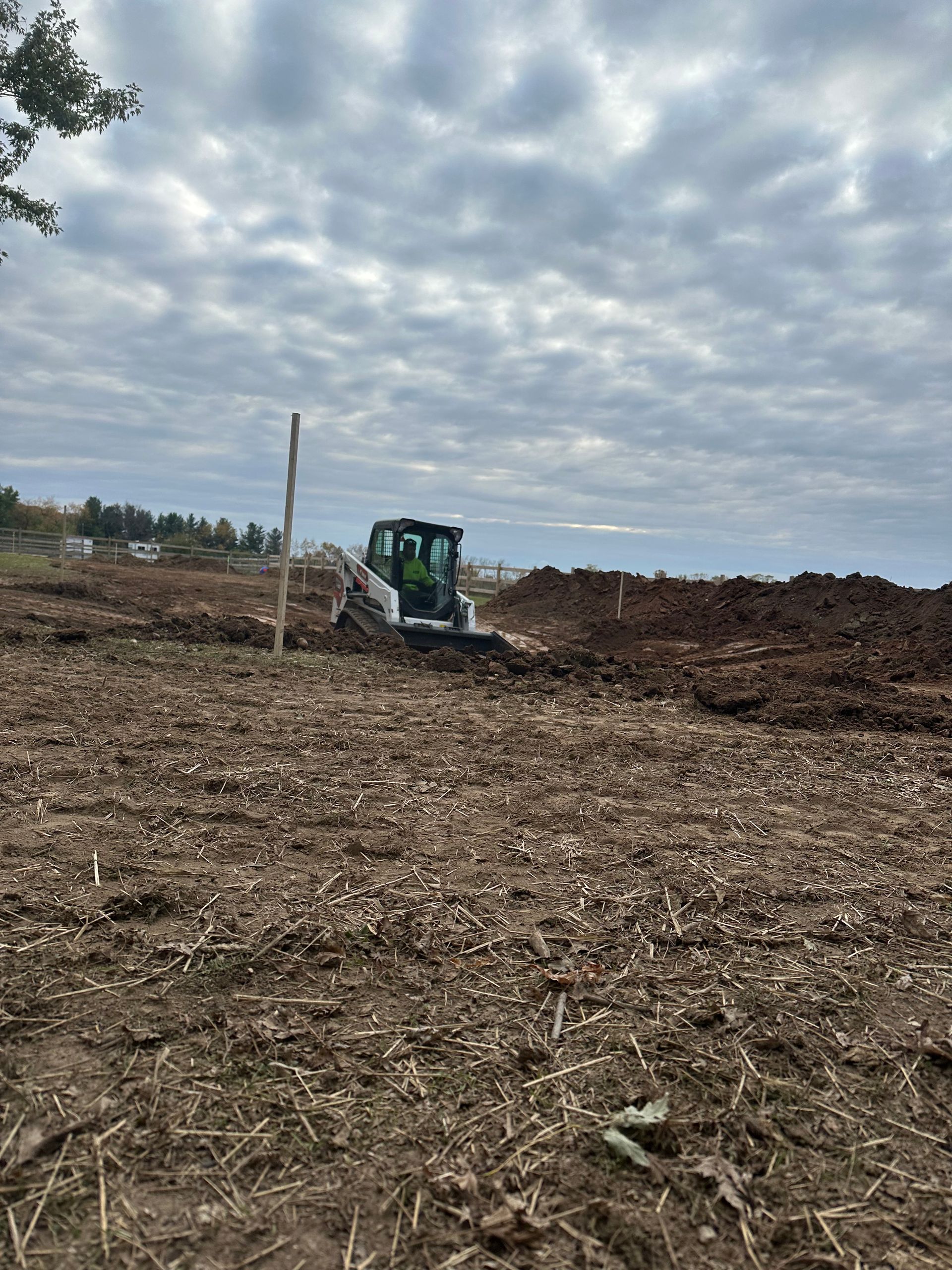 A white skid-steer loader works in an open, dirt-covered field under a cloudy sky.