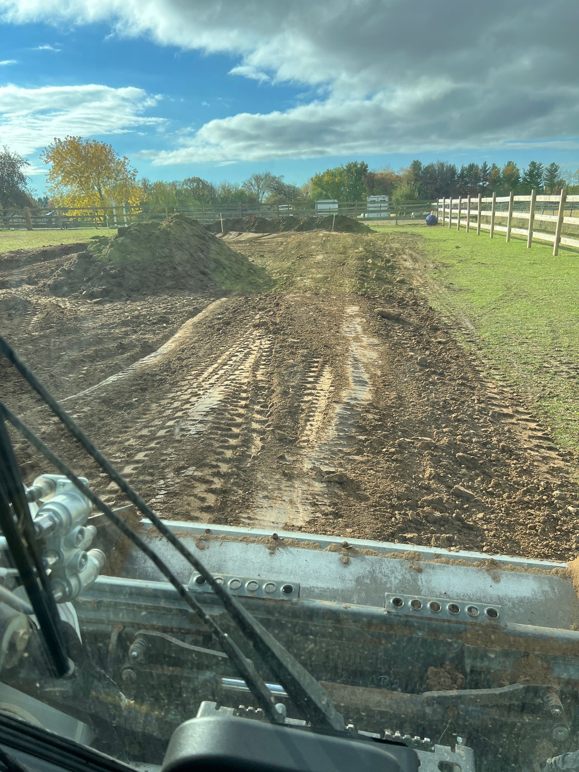 View from inside heavy machinery looking out at a freshly cleared, muddy field next to a wooden fence on a sunny day.