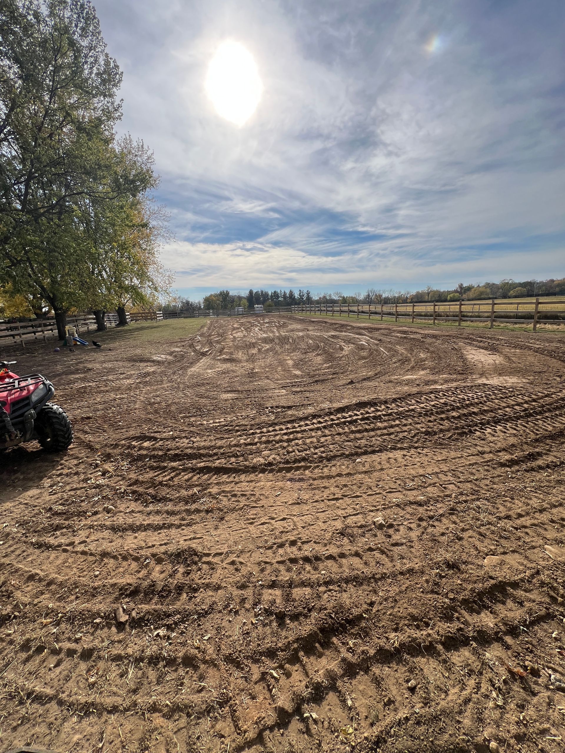 A sunlit, tilled field showing fresh tire tracks in the dirt, with trees on the left and a fence line in the distance.