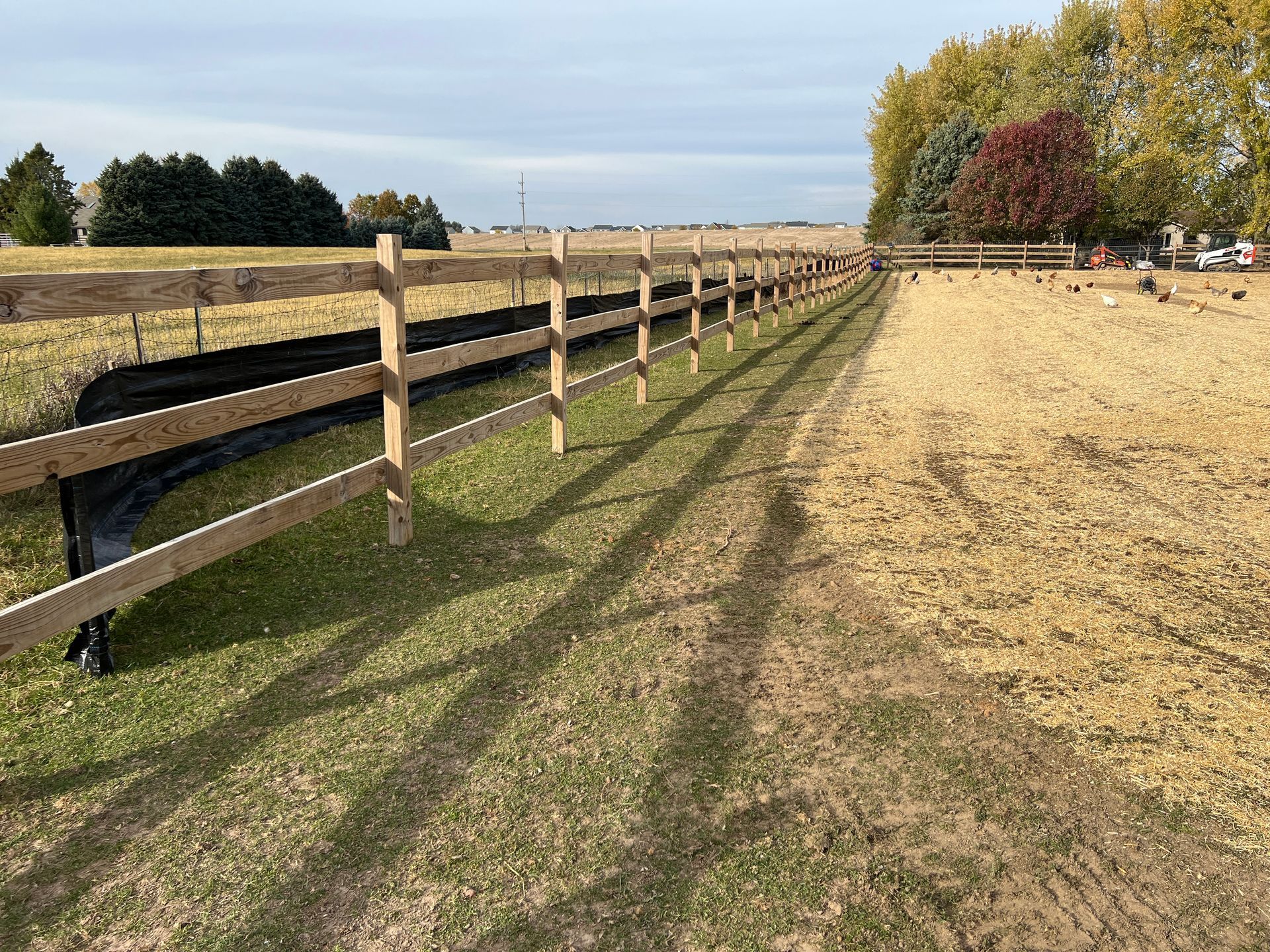 A wooden post-and-rail fence runs through a grassy field, with a black silt fence installed along its base.