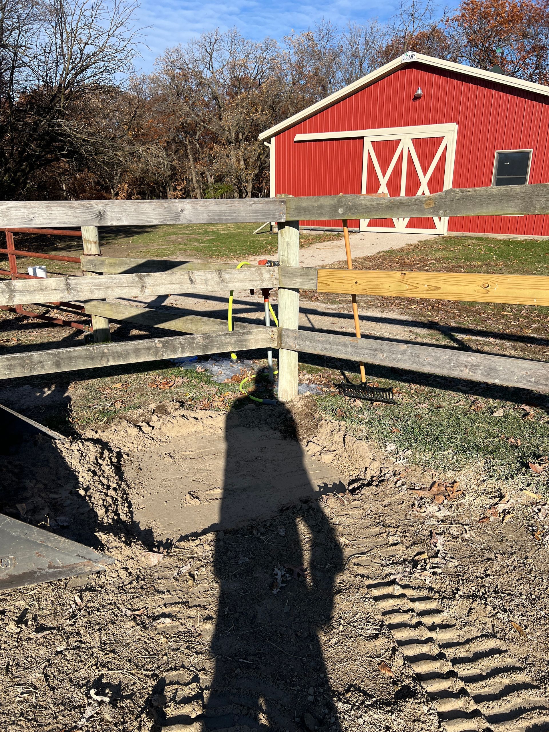 A red barn sits behind a wooden fence, with a person's long shadow cast on the gravel ground in the foreground.