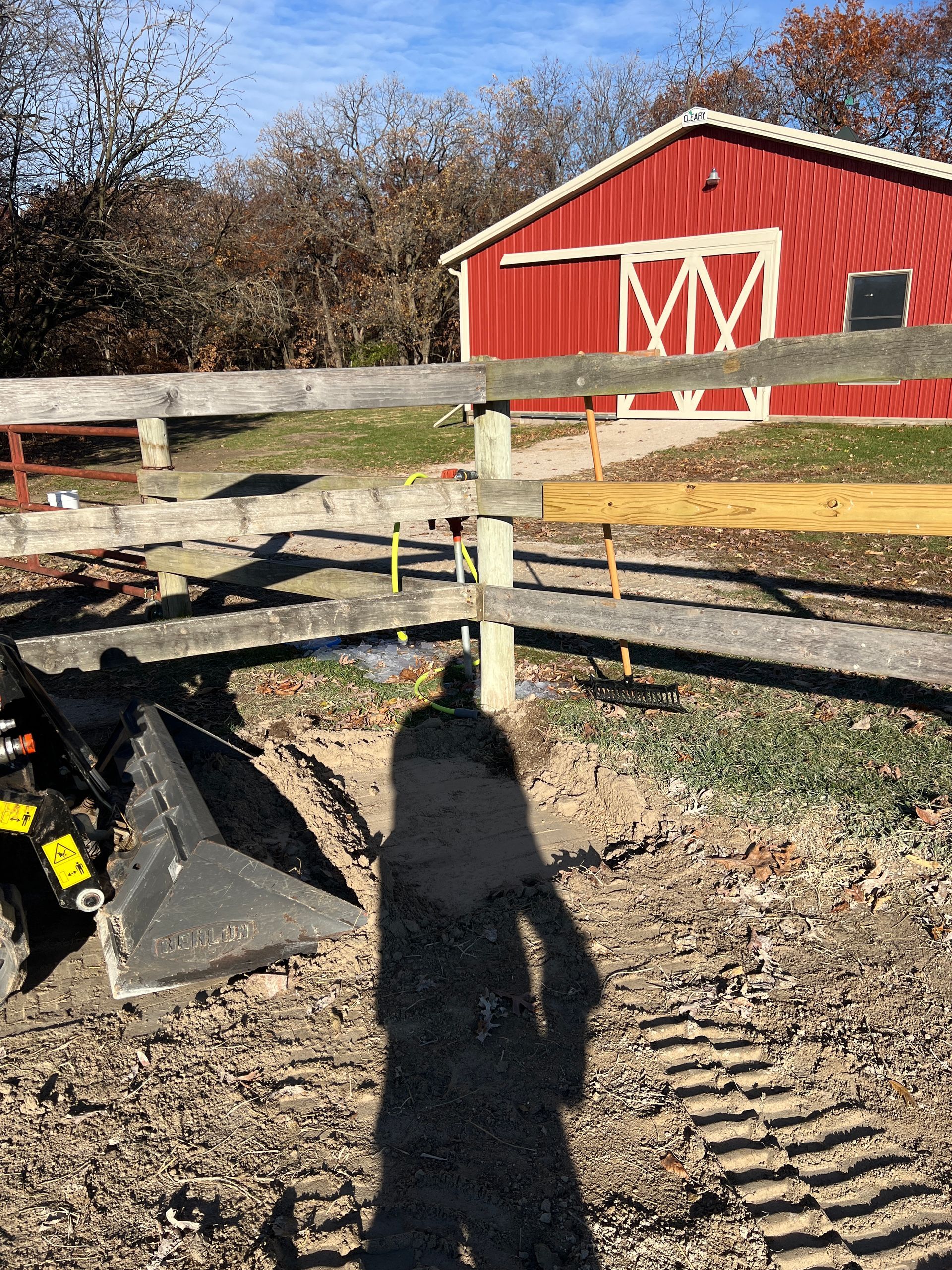A metal tractor attachment sits on dirt near a wooden fence, with a red barn in the background under a blue sky.