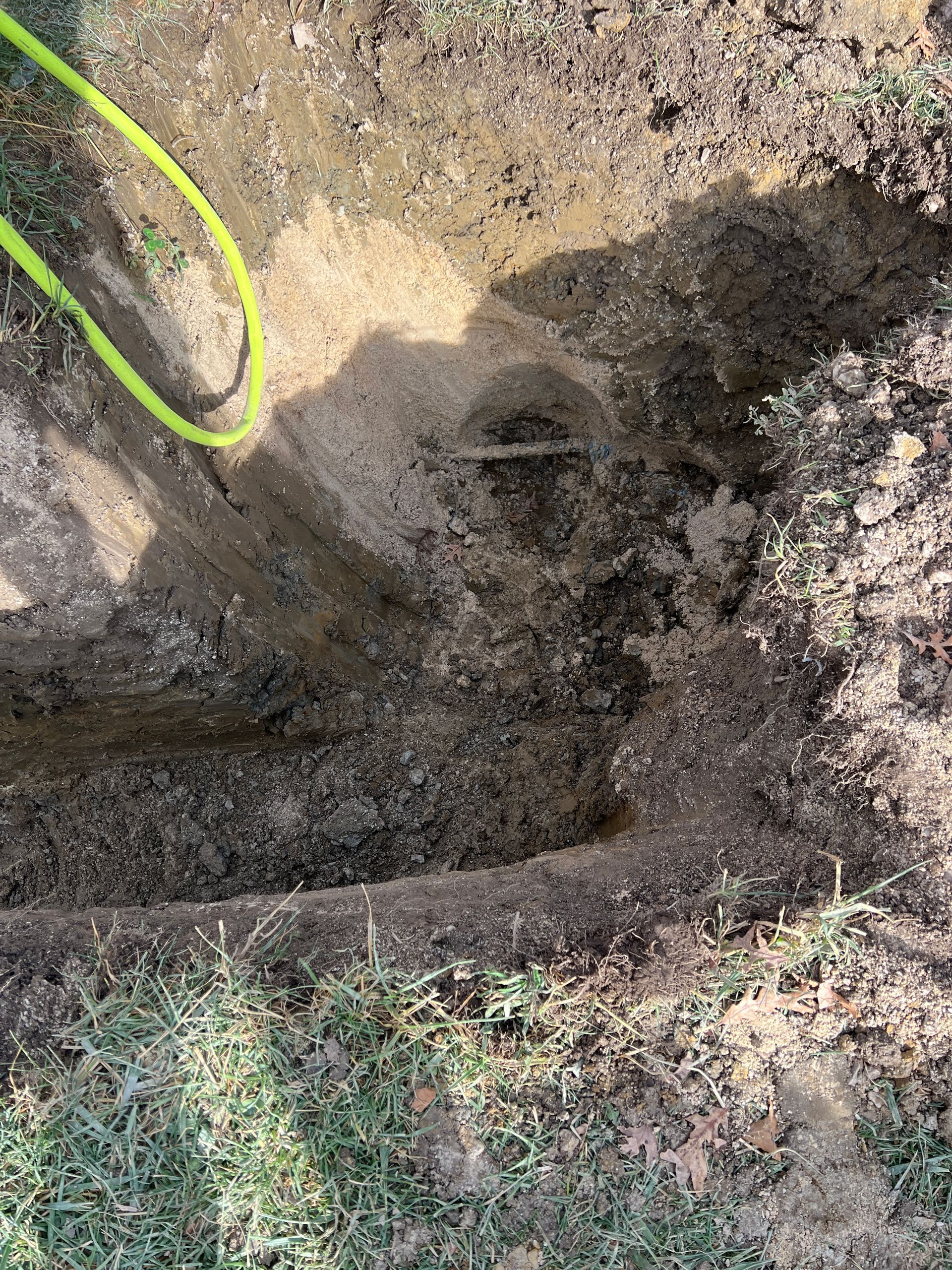 An excavated dirt pit in a grassy yard, showing an exposed utility pipe and a bright green hose extending into the hole.