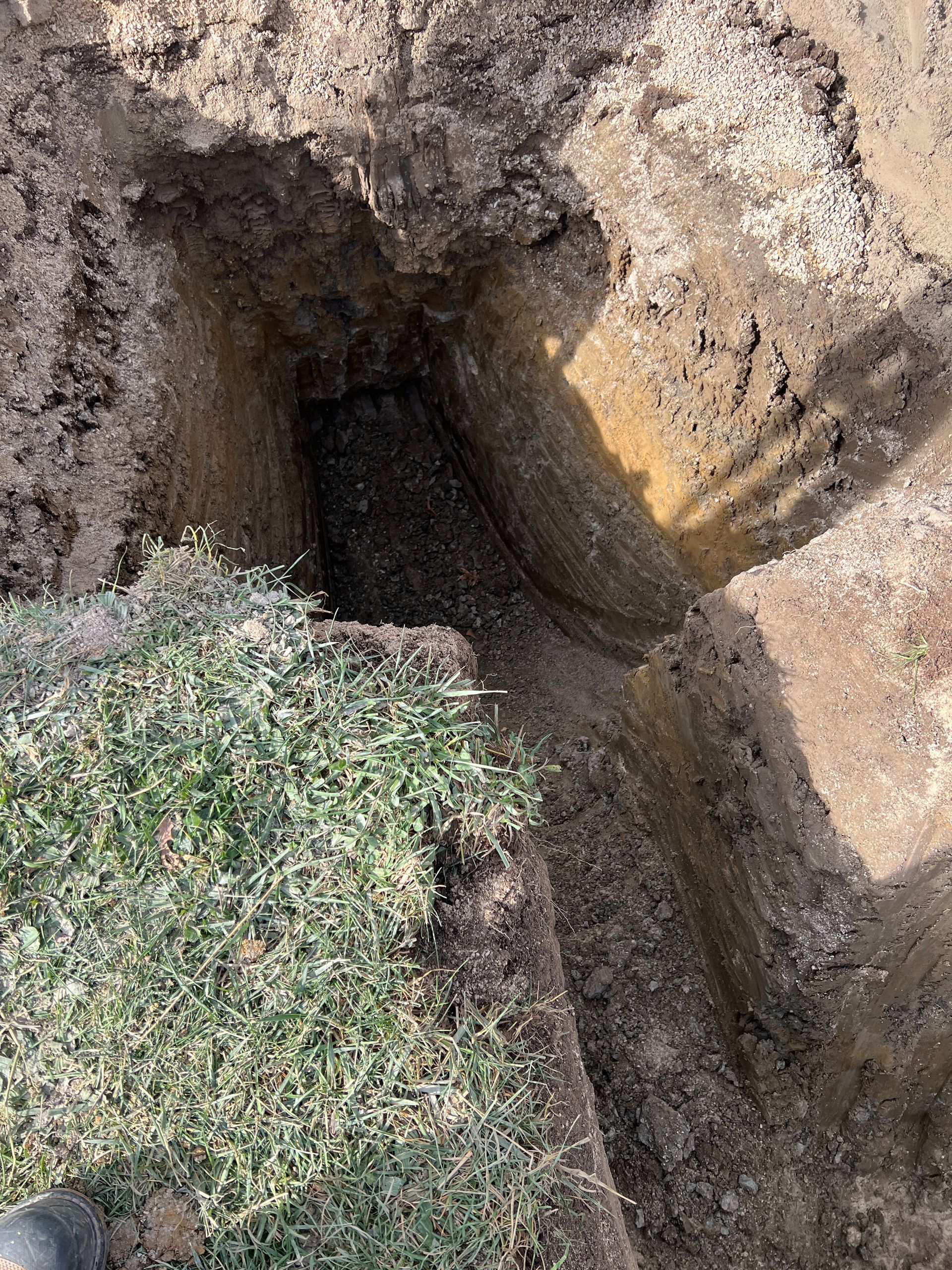 A vertical, rectangular hole dug into the ground next to a patch of green grass, showing layered soil in the pit.