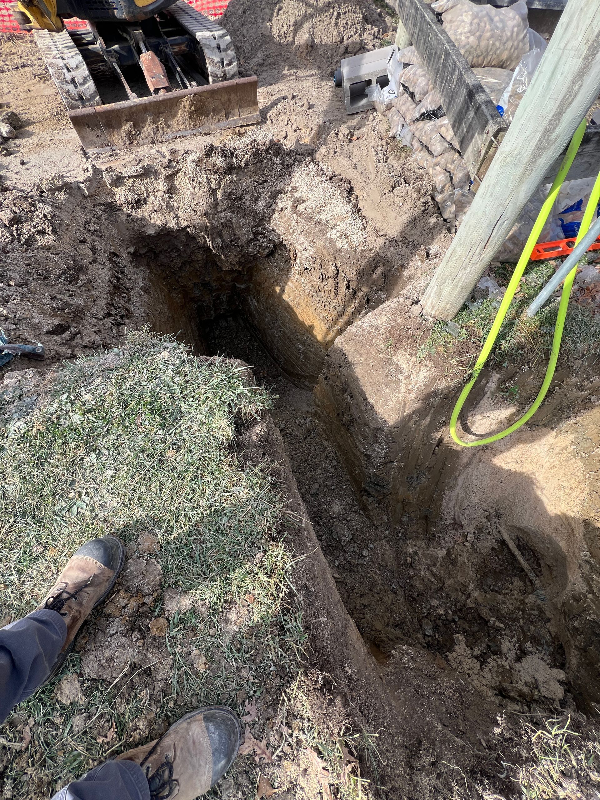 A view from above showing a deep, narrow trench dug into soil next to a small excavator and a bright yellow strap.