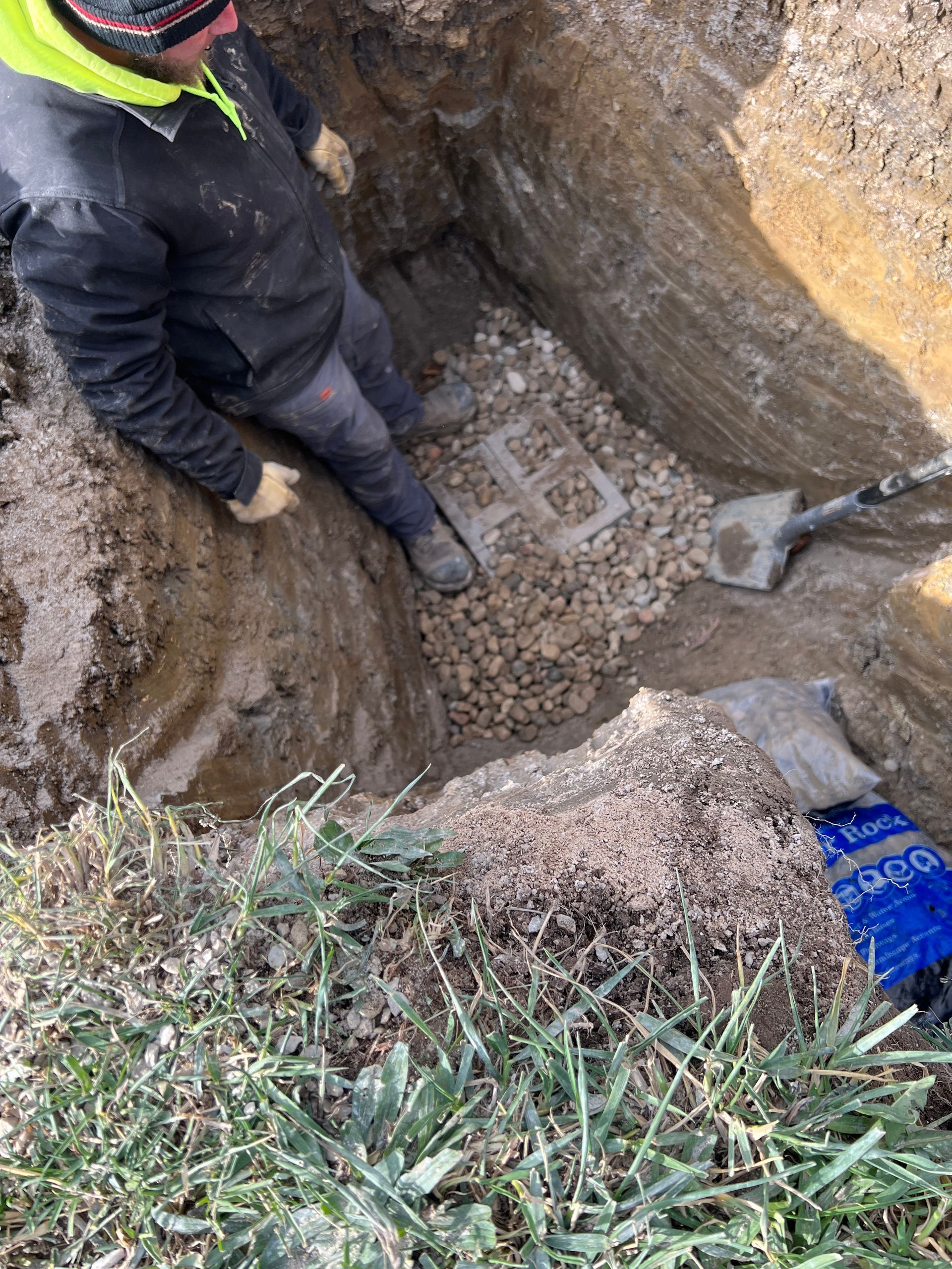 A person in a high-visibility jacket stands inside a deep, soil-filled excavation pit next to a shovel and buried pipe.
