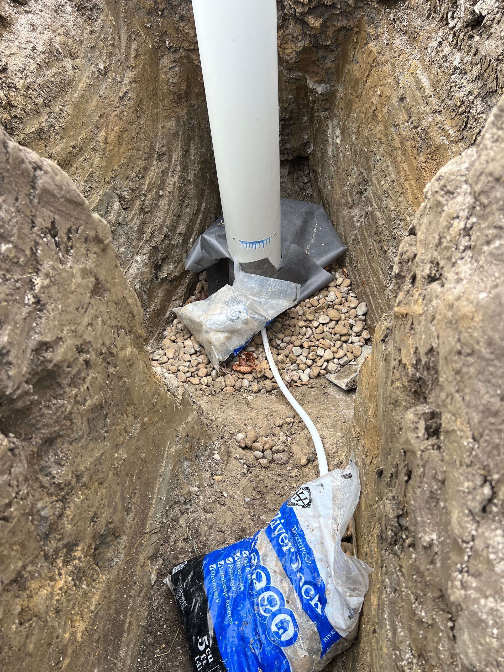 A white vertical pipe set in a narrow dirt trench, with gravel and a partially buried bag of construction material.