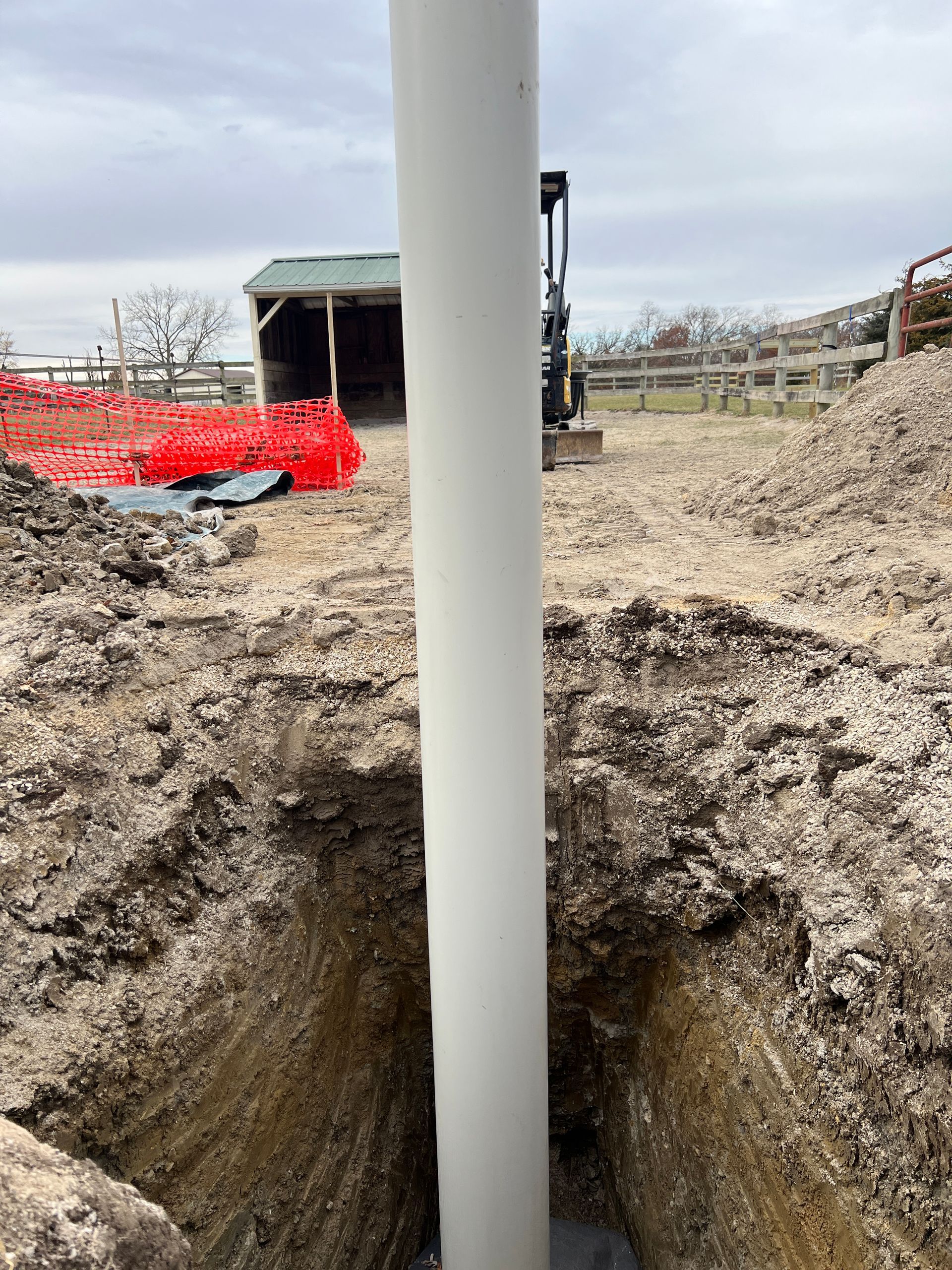 A vertical white pipe stands in an excavated dirt trench at a construction site with a shed and heavy machinery in the back.