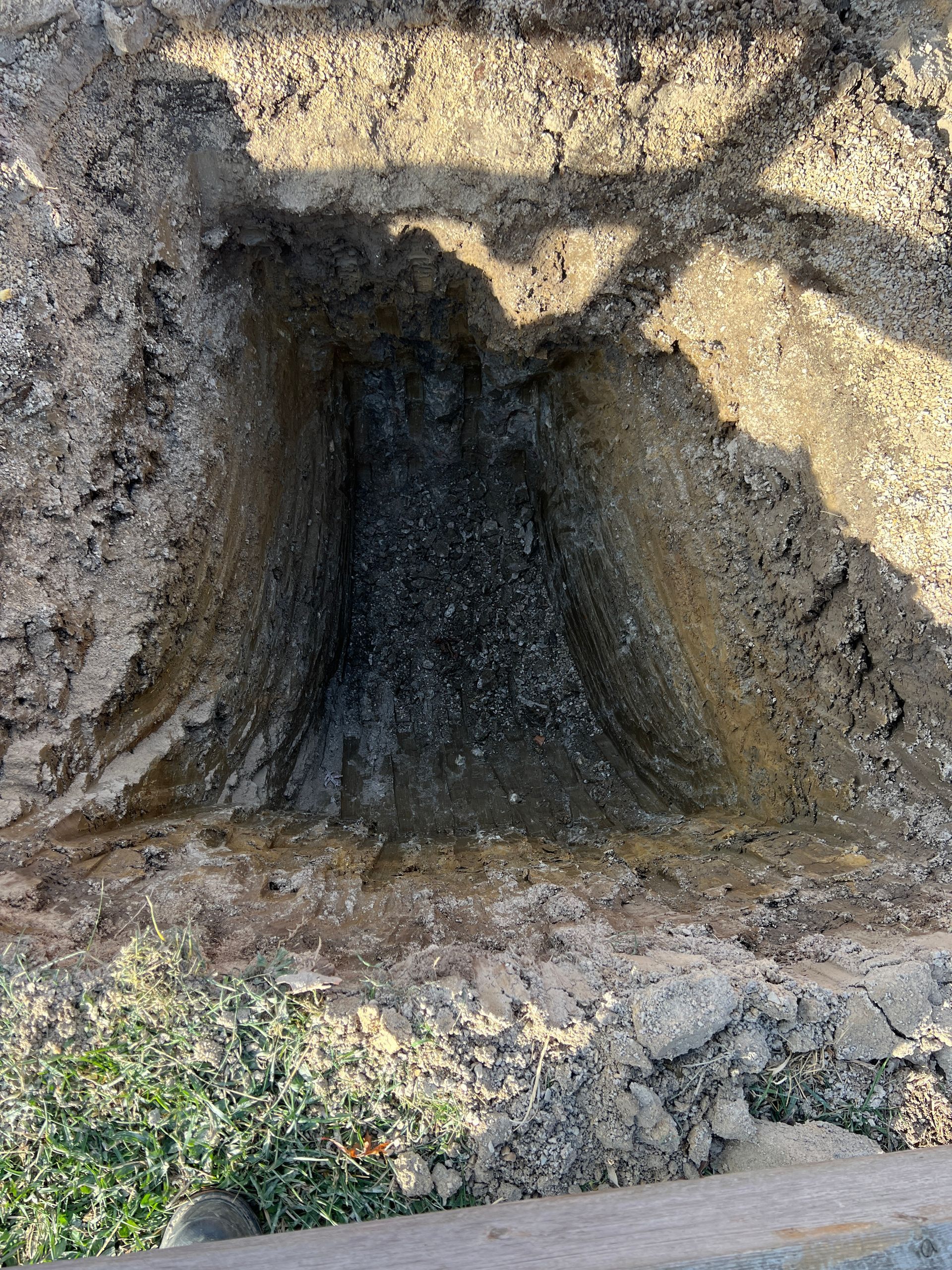 A deep, rectangular pit dug into the ground, revealing layers of dark soil and earth with a shadowy cast across the top.