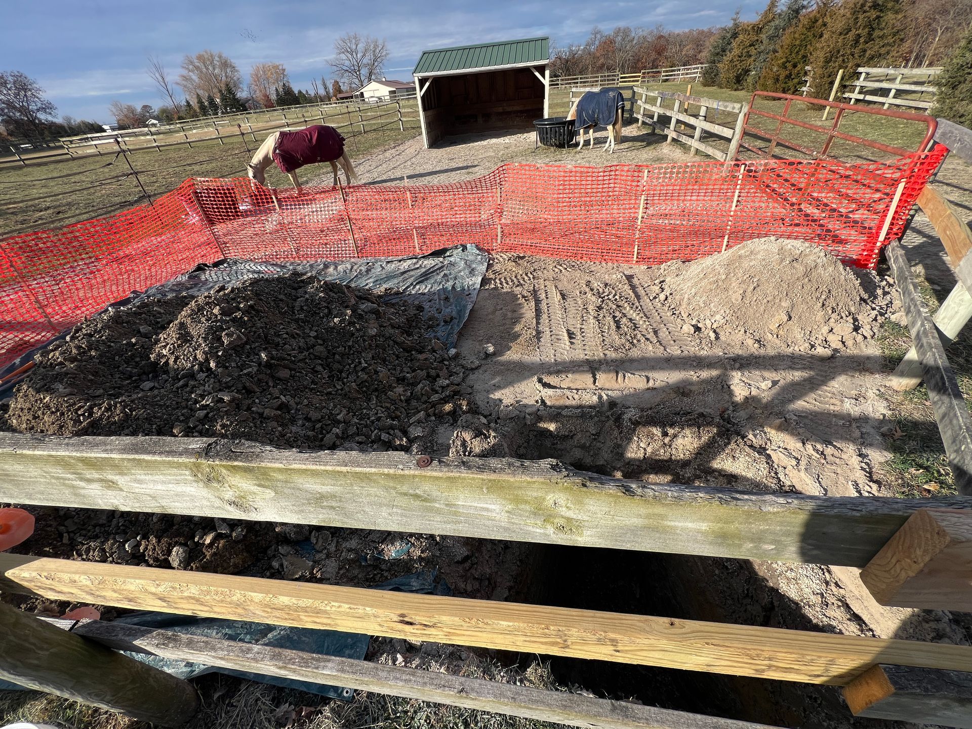 Two horses in blankets graze in a fenced area containing a large pile of soil and mulch next to an open excavation.