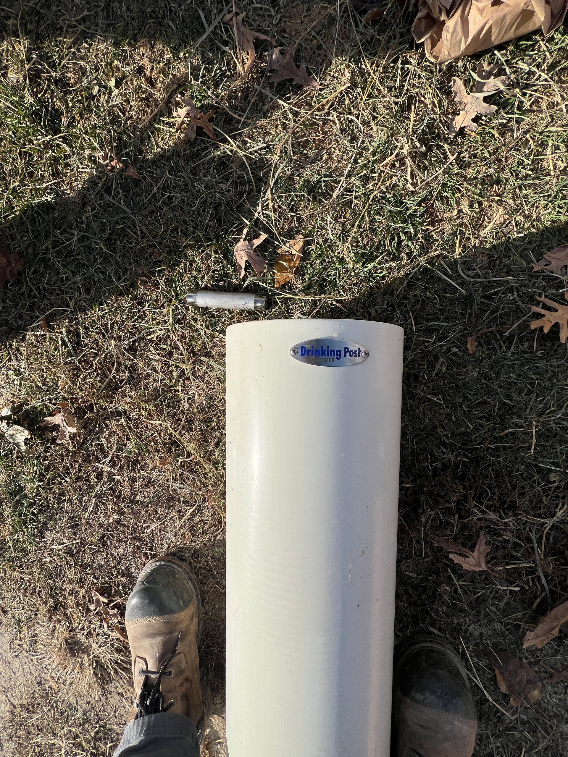 A cylindrical, white Stratos-brand pipe component sits on a patch of grass next to a pair of work boots.