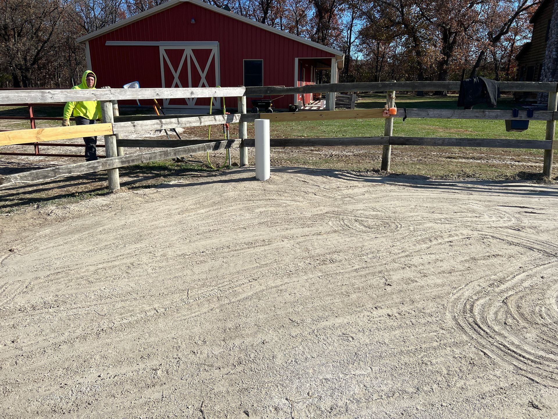 A person in a neon yellow jacket stands by a wooden fence in a gravel outdoor arena near a red barn.
