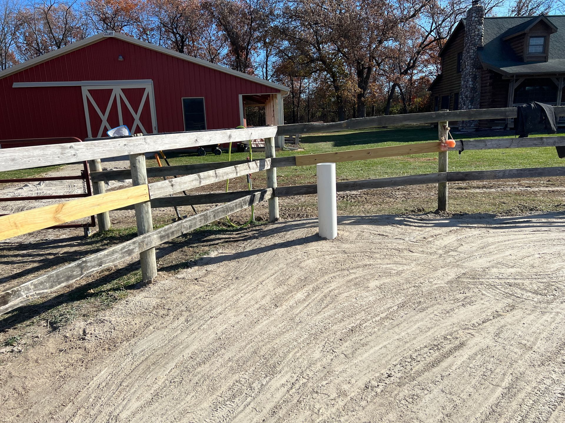 A red barn and a log cabin stand behind a wooden fence enclosing a sandy training ring with a white post in the center.