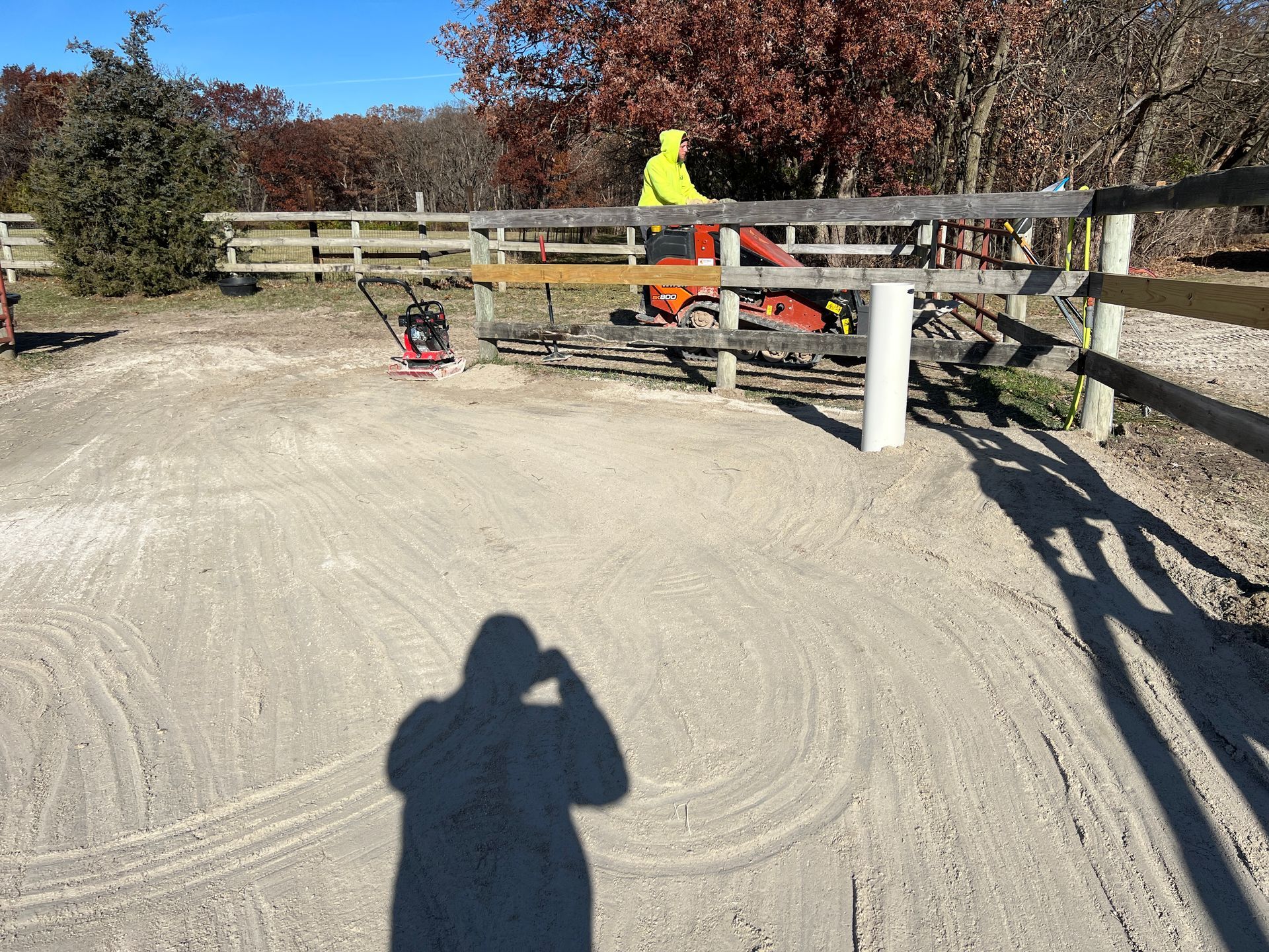 A person in a neon yellow jacket operates a riding mower near a wooden fence bordering a sandy, graveled outdoor area.
