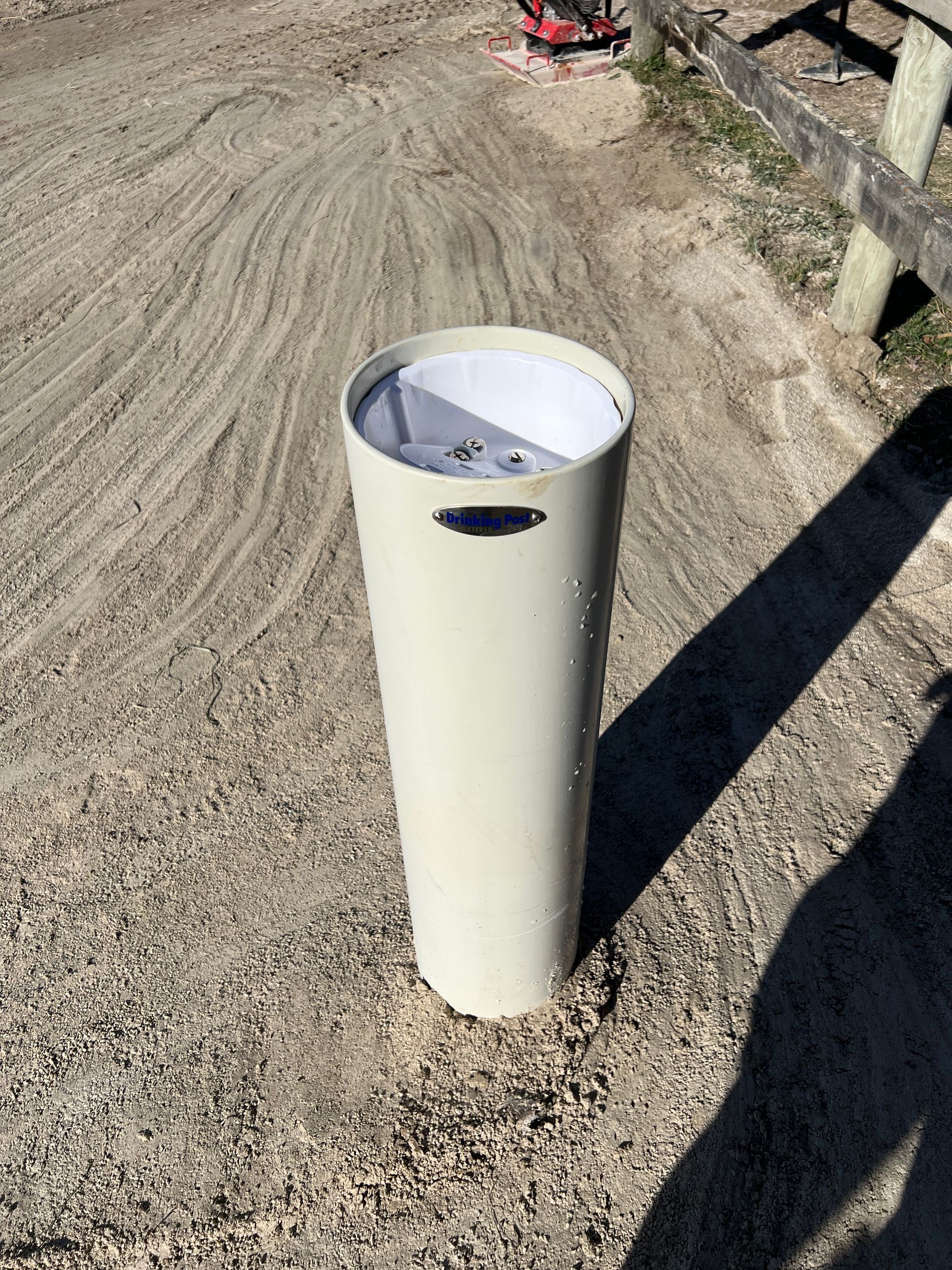 A beige, cylindrical bollard with a blue label stands upright in a gravel area next to a wooden fence.