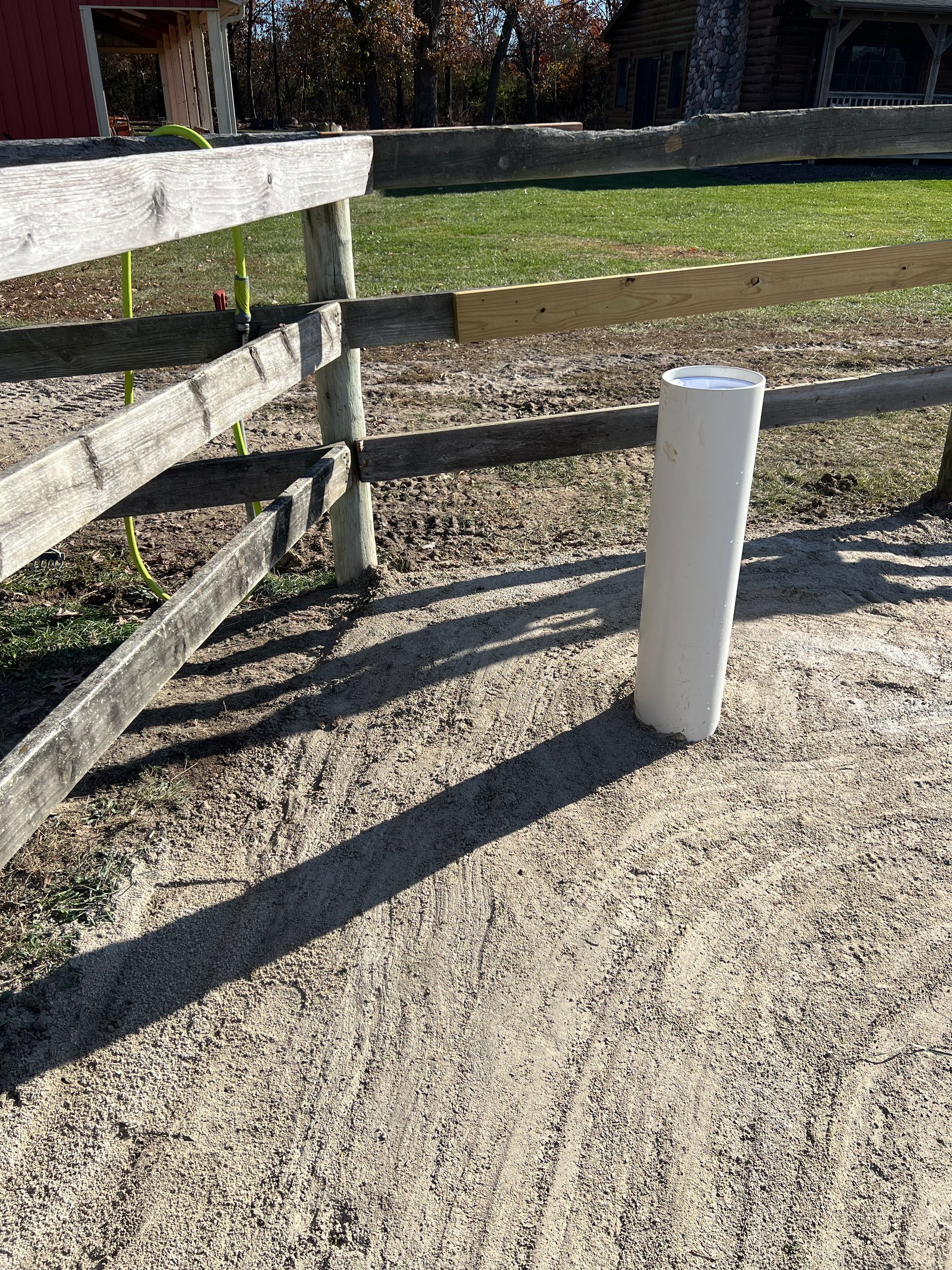 A white PVC pipe stands vertically in a gravel area next to a weathered wooden post-and-rail fence.