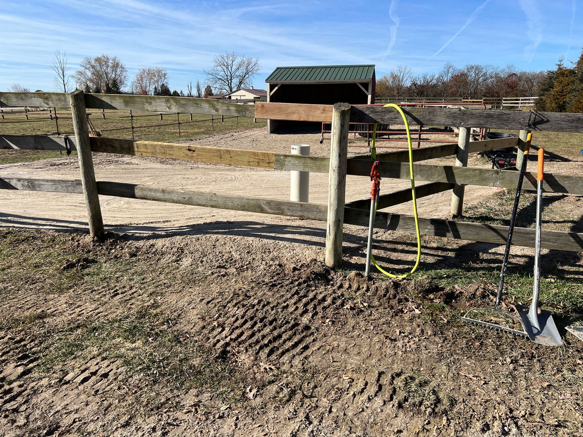 A wooden fence surrounds a dirt paddock containing a small, green-roofed shed, with a shovel leaning against the fence.
