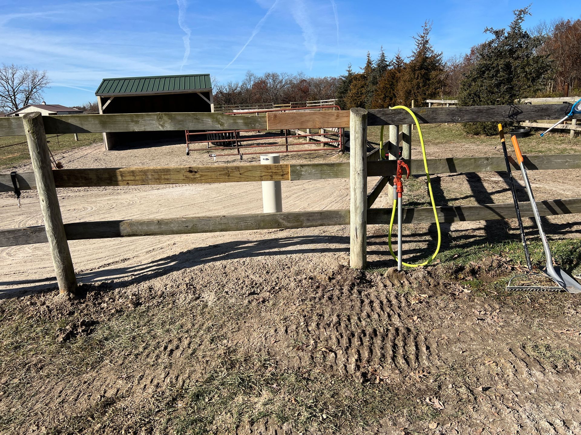A wooden post-and-rail fence surrounds a sandy paddock with a green shed in the background under a clear blue sky.
