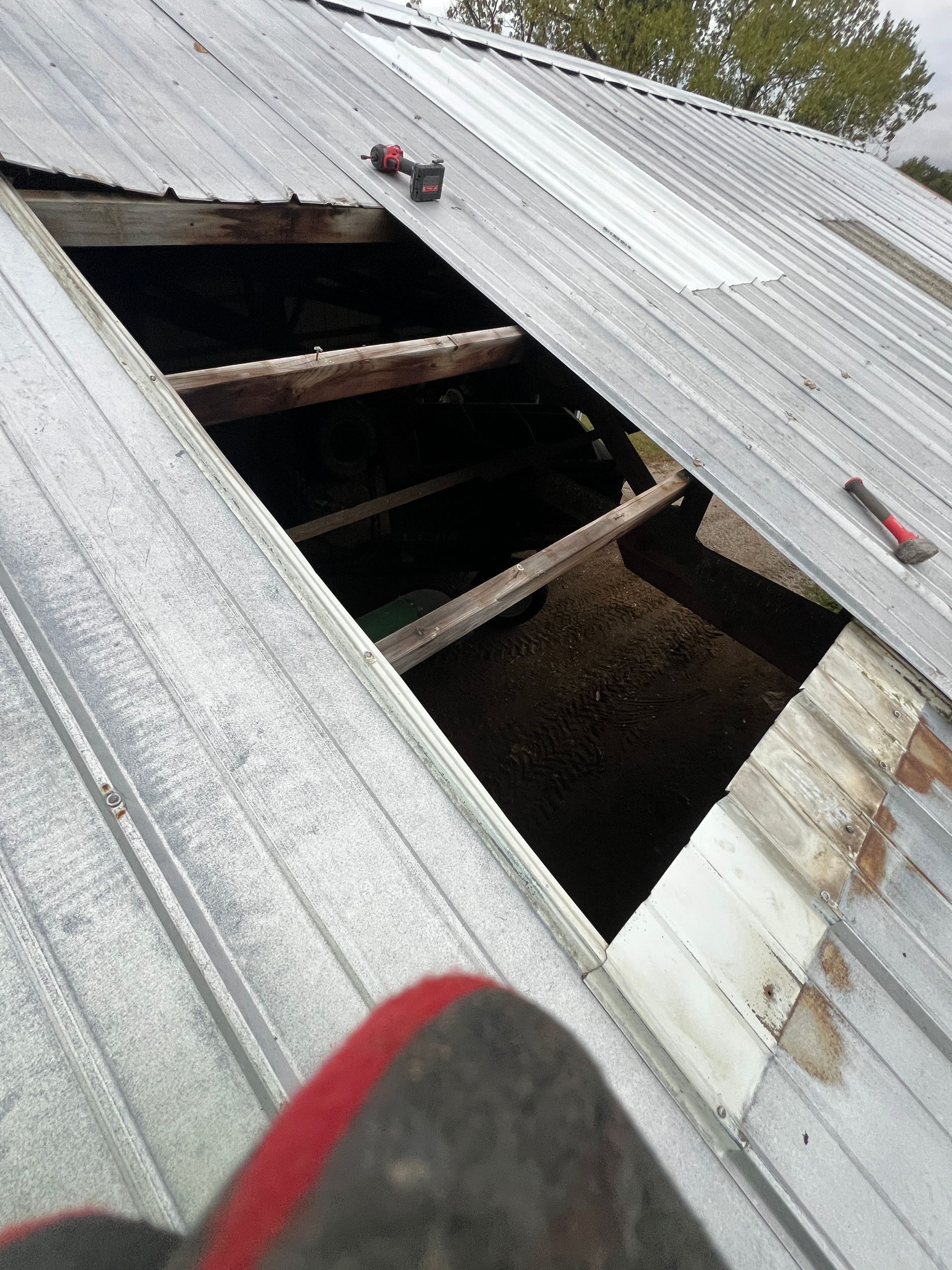 A view from a roof looking down through a large hole where metal panels are missing, revealing wooden rafters and dirt.