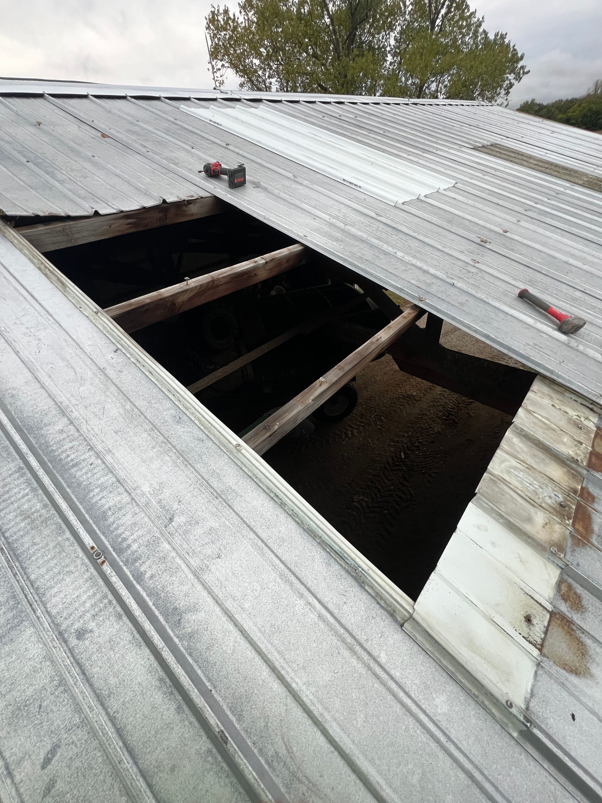 A large rectangular hole in a corrugated metal barn roof, exposing the dark interior and wooden support beams below.