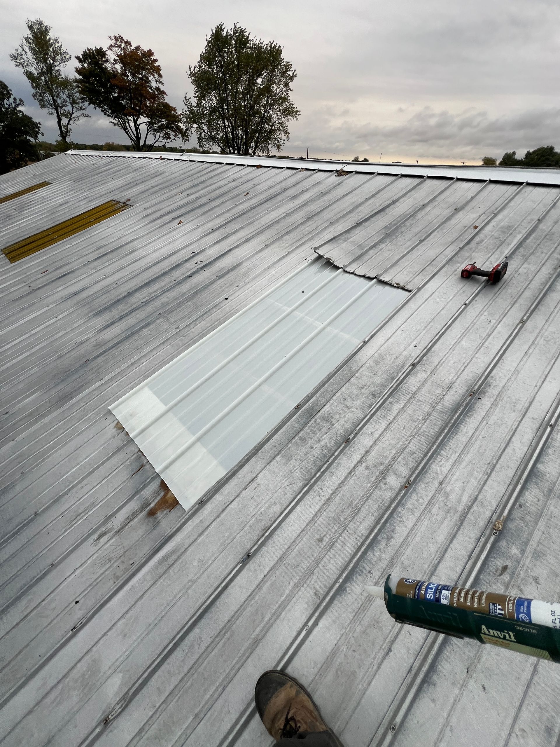 A person standing on a metal roof, installing a translucent skylight panel with a caulk gun nearby.