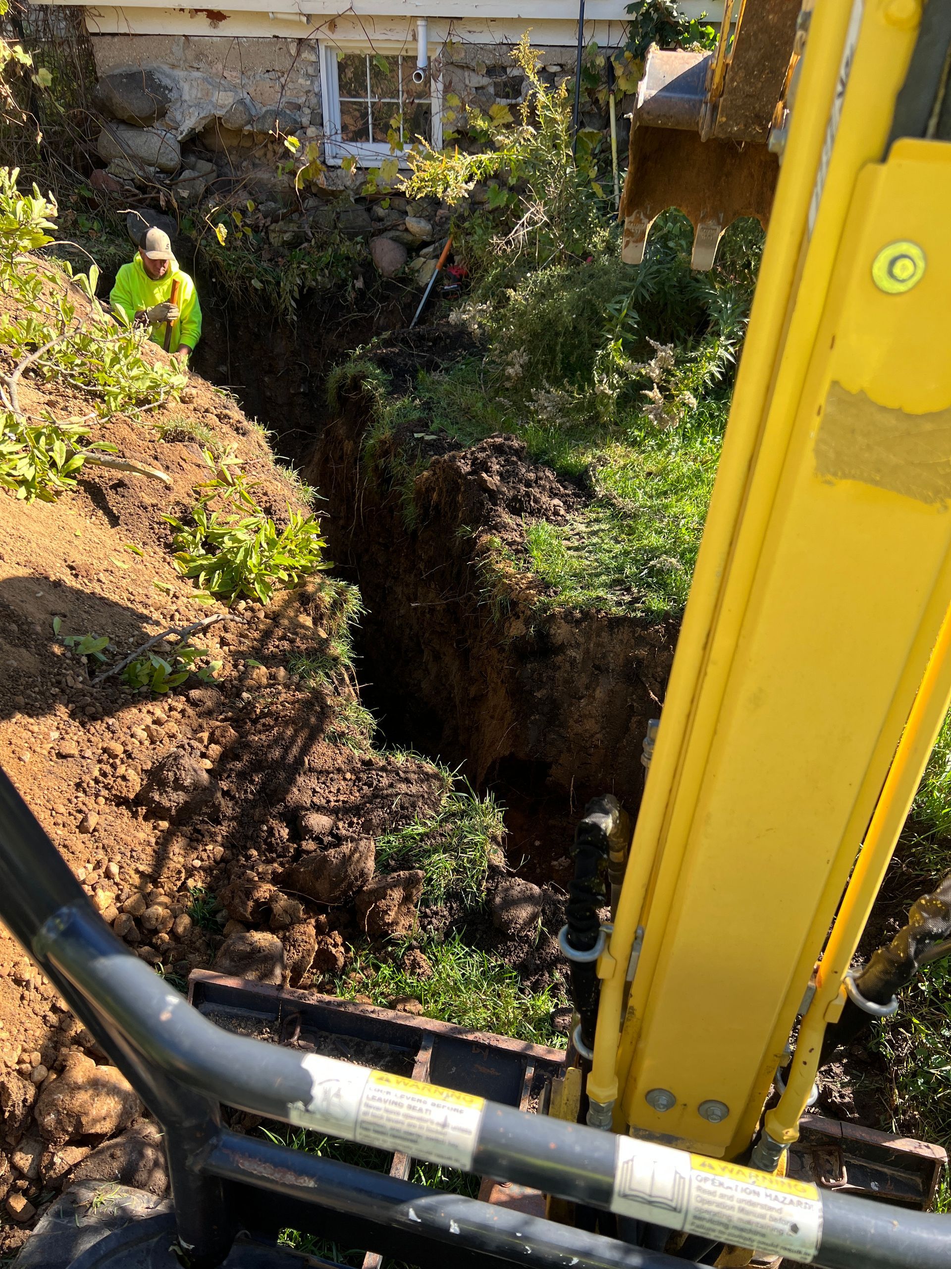 A worker in a bright yellow vest stands in a deep trench next to a house foundation, seen from an excavator's perspective.