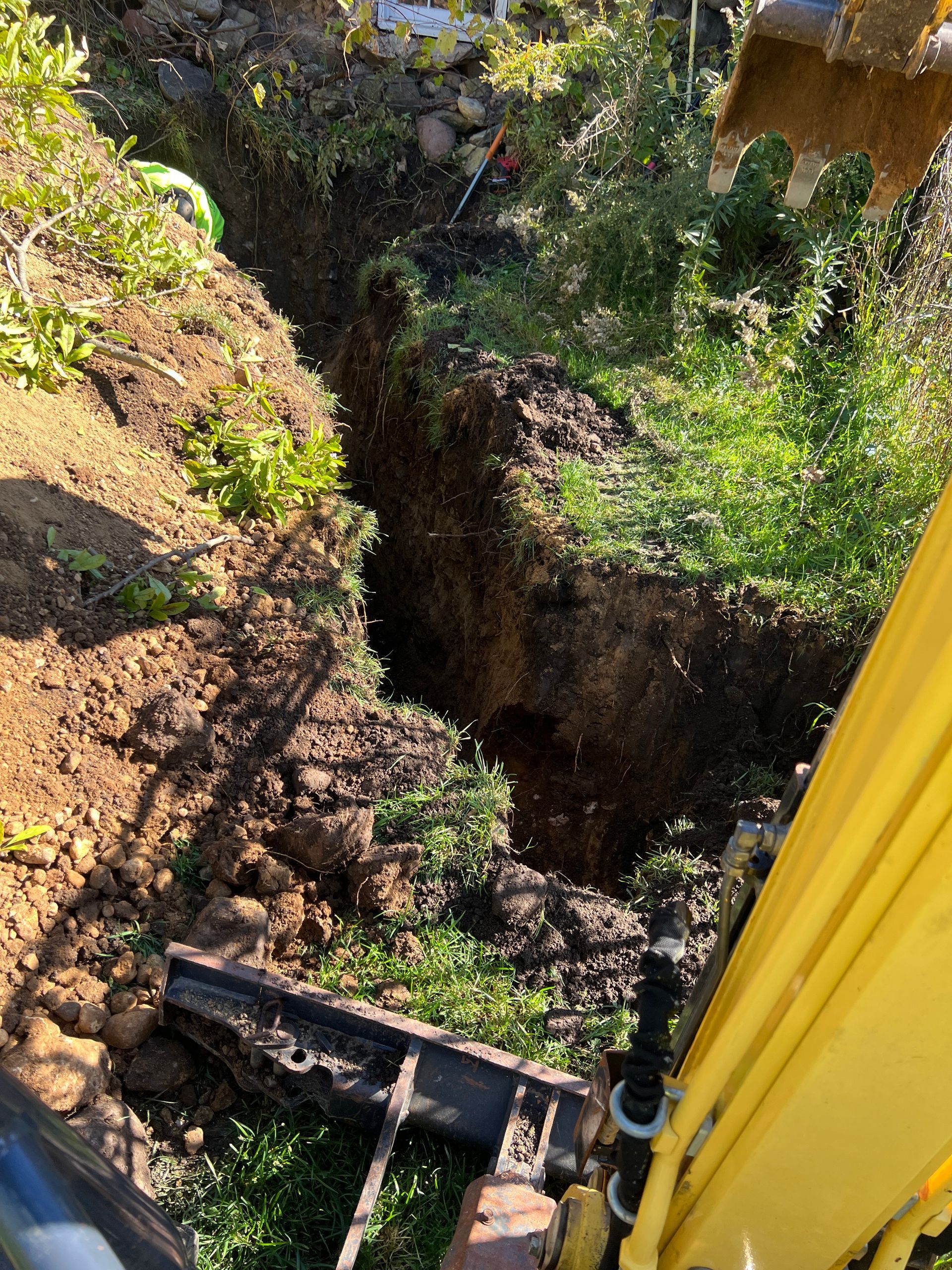 View from an excavator cockpit overlooking a deep, narrow trench dug into an earthen hillside covered with vegetation.