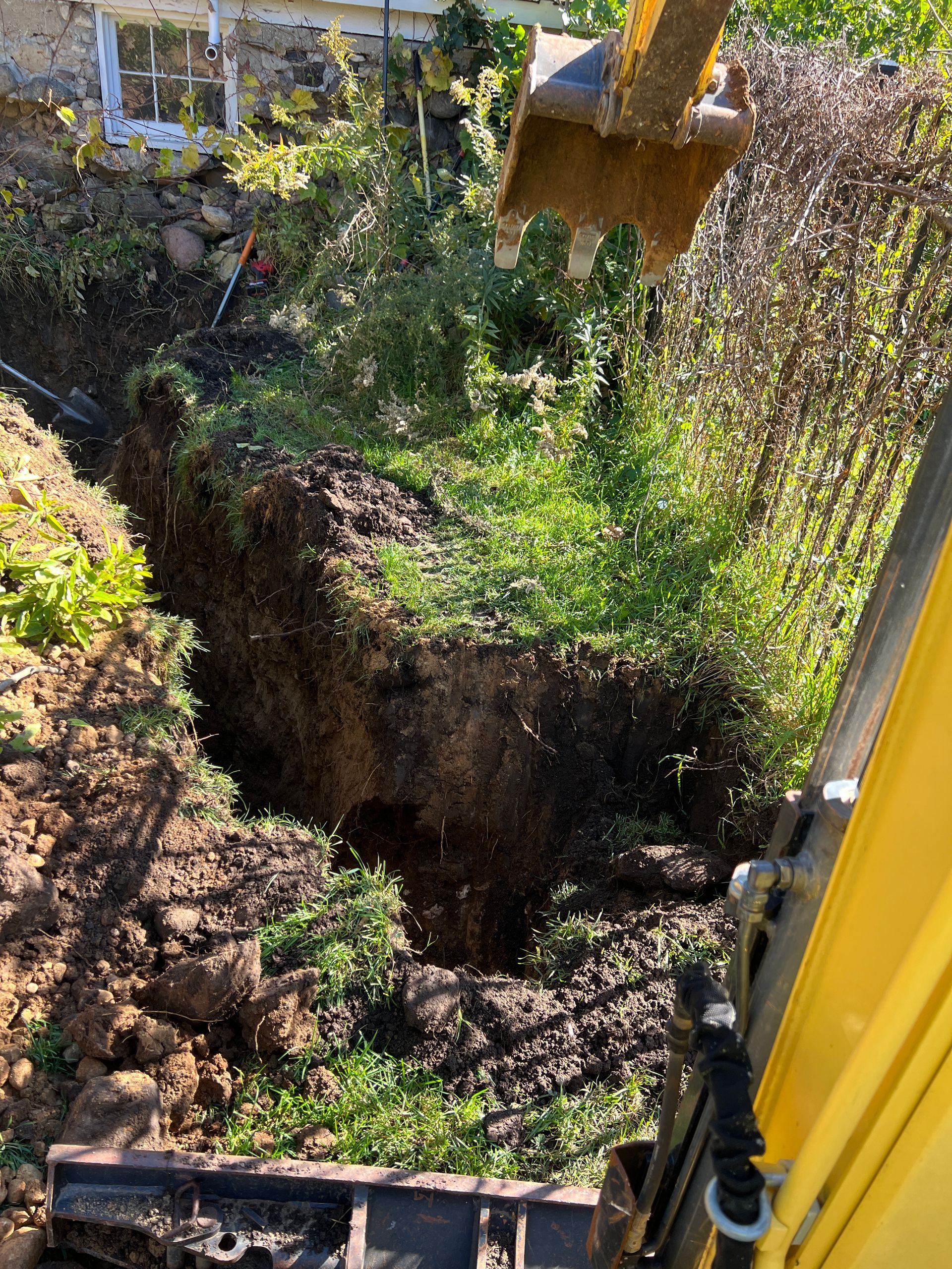 View from an excavator cab looking down at a hole being dug in a grassy yard next to a stone building foundation.