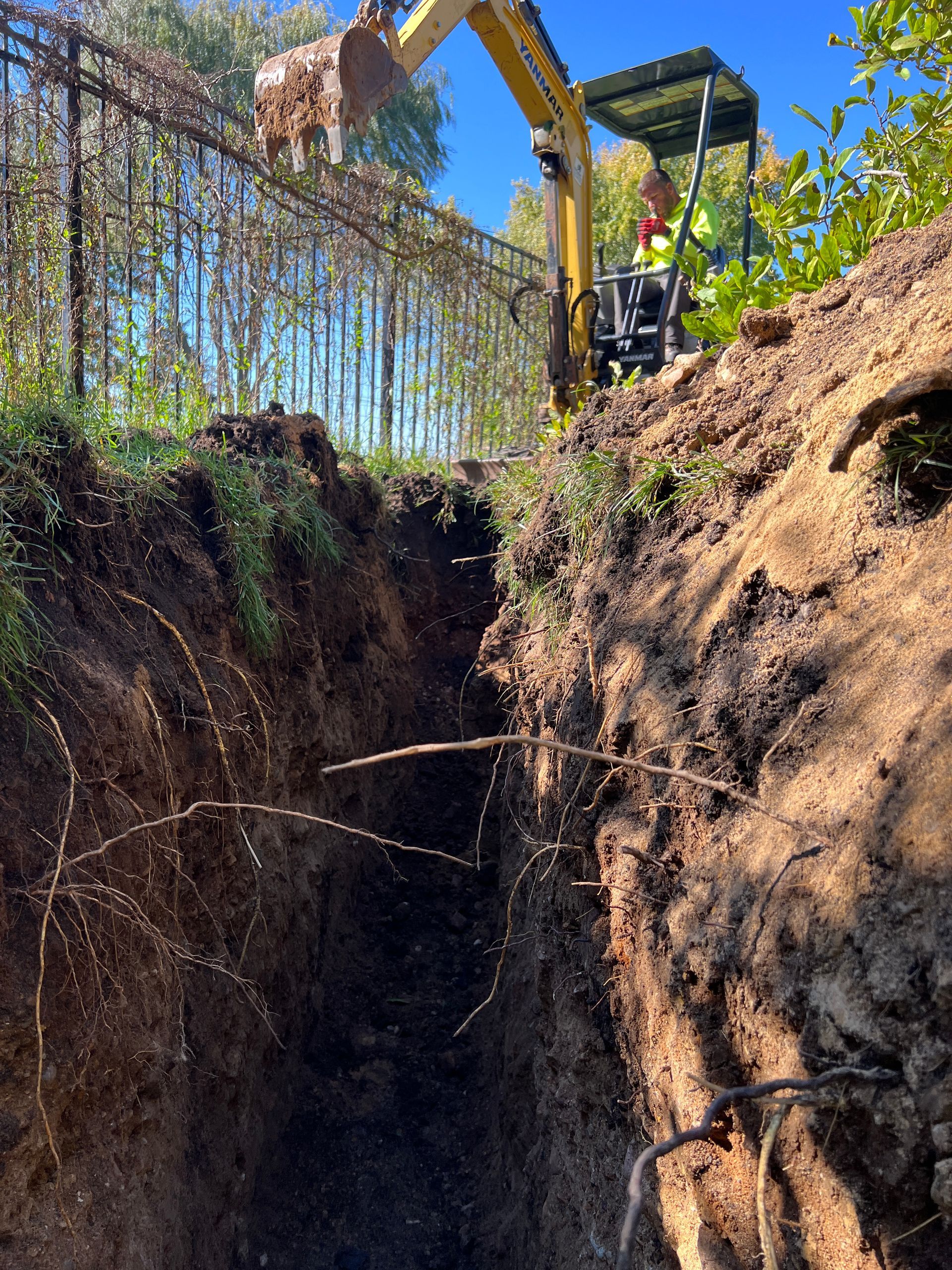 A yellow excavator digging a deep trench in the ground alongside a fenced area on a sunny day.