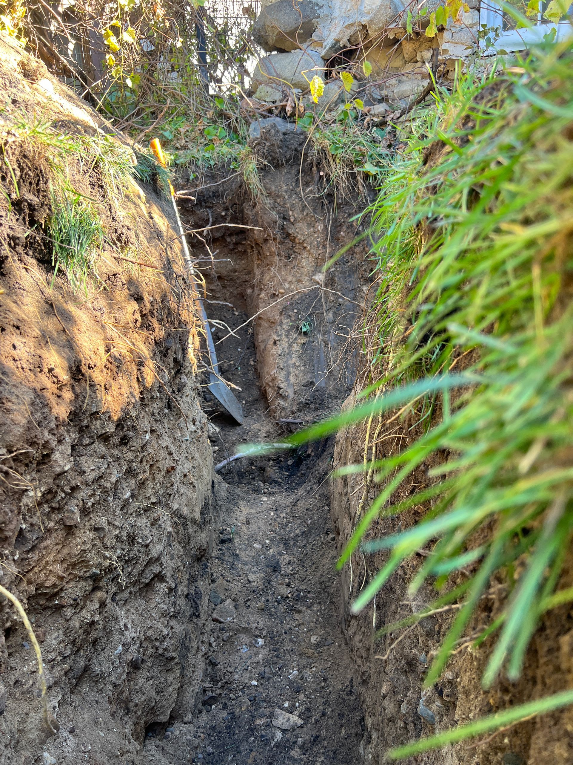 A narrow, deep trench dug into the earth, with a shovel standing inside and green grass bordering the top edge.