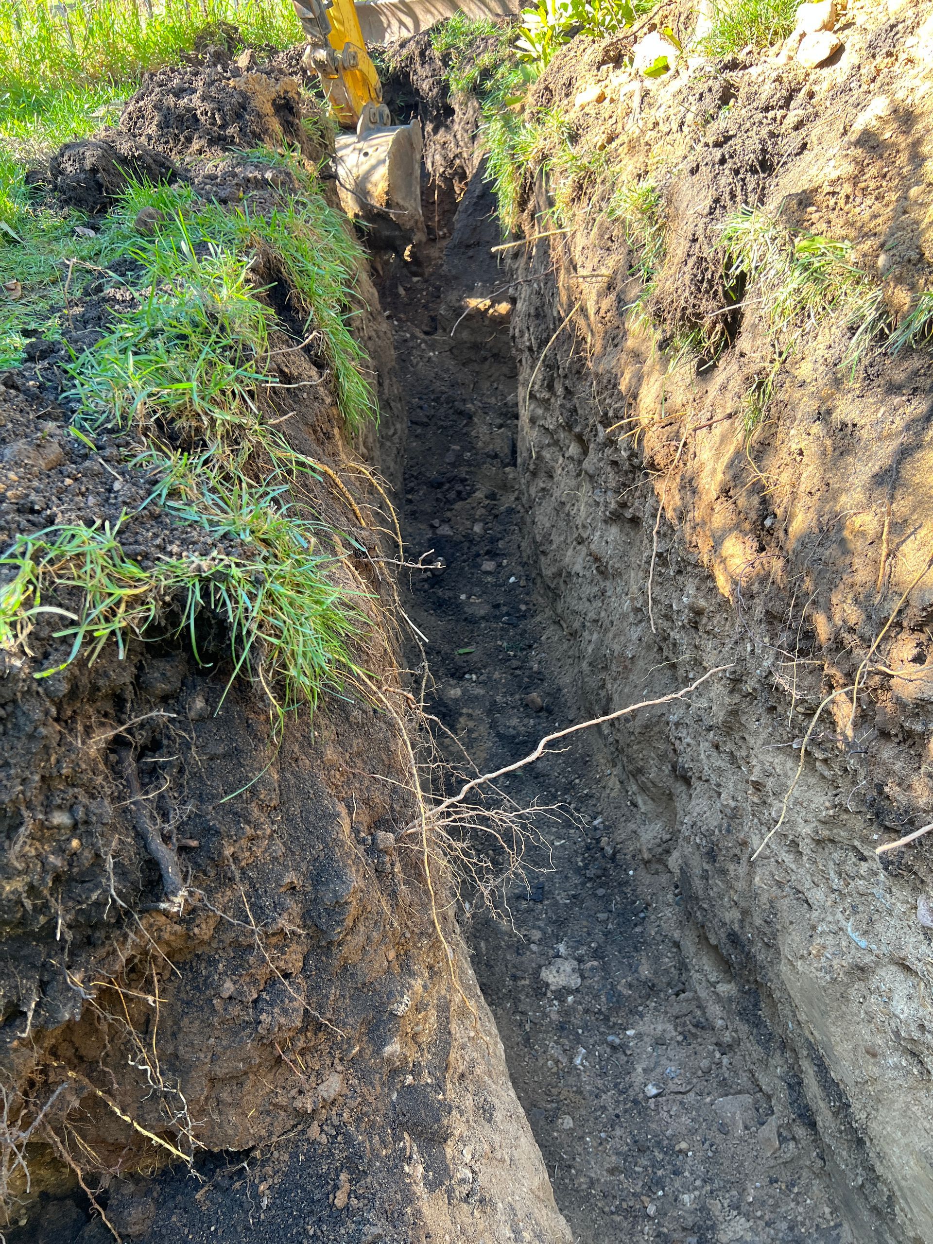 A narrow, deep trench dug into dark soil, bordered by grass and sunlight, with a glimpse of yellow machinery at the end.
