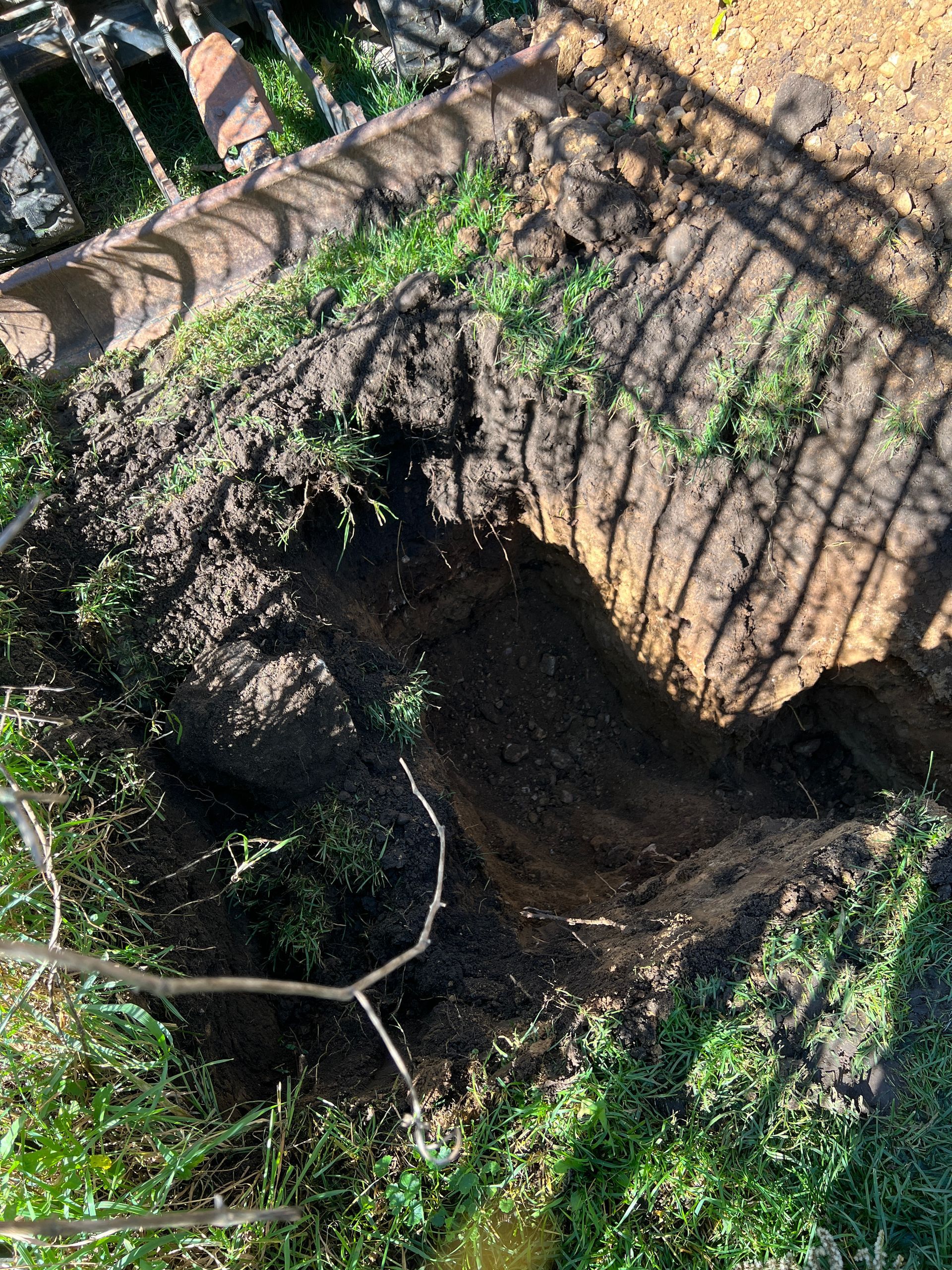 A deep, rectangular pit dug into the ground, surrounded by grass and dirt, partially cast in shadow.